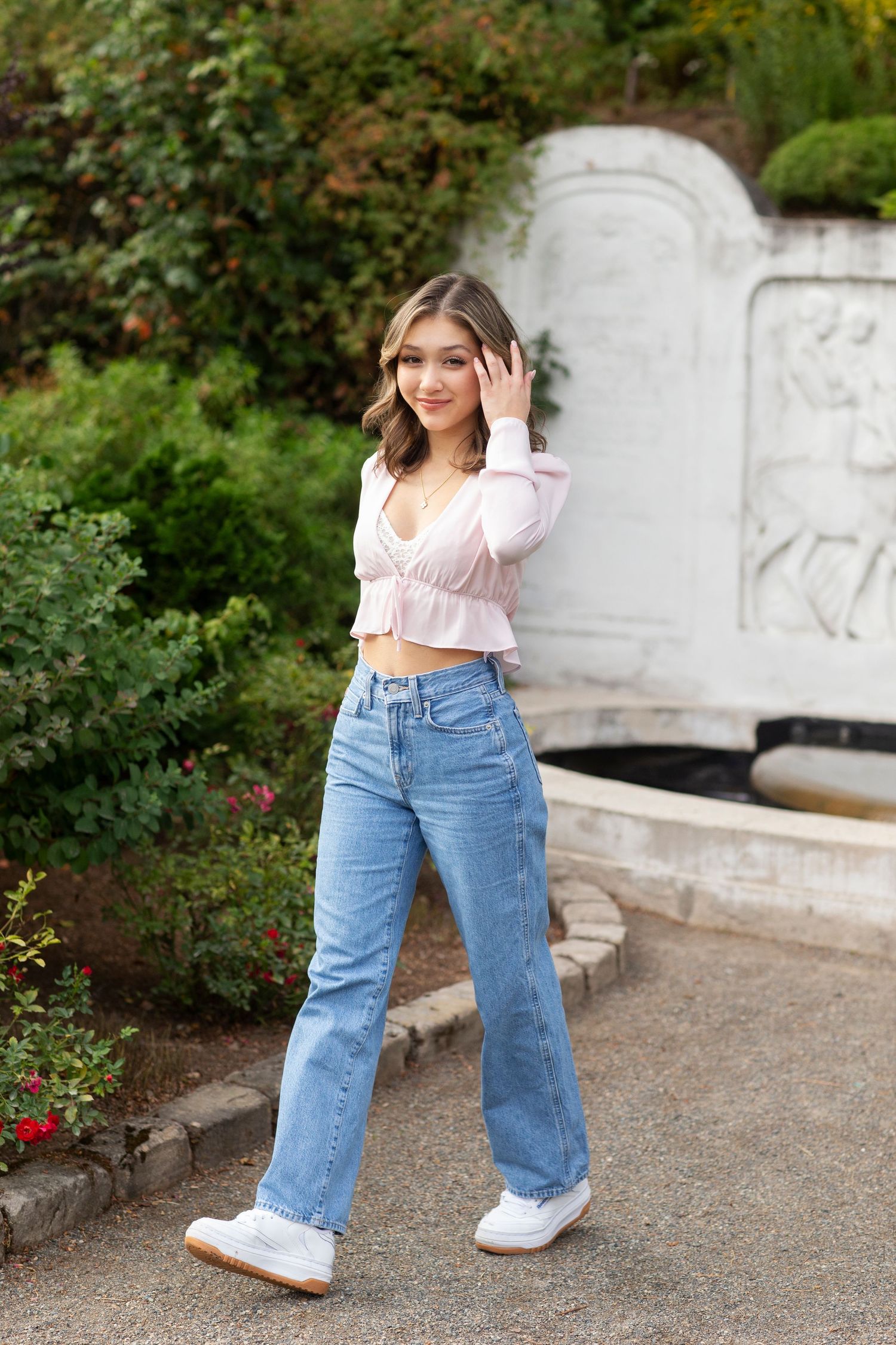 Young woman in pink crop top and jeans walking near a decorative stone fountain in a garden.