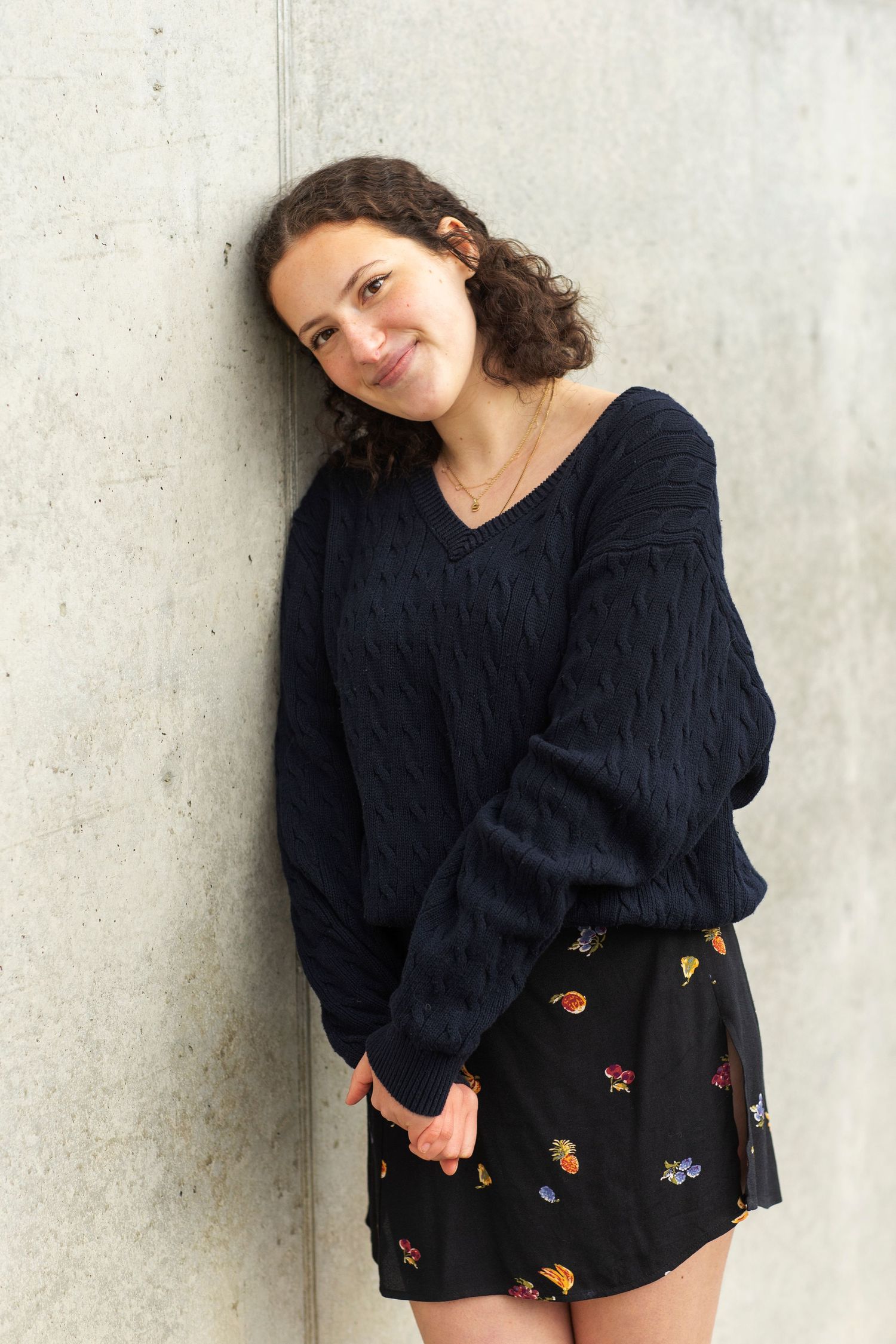 Young woman with curly hair in navy sweater and floral skirt leaning against concrete wall.
