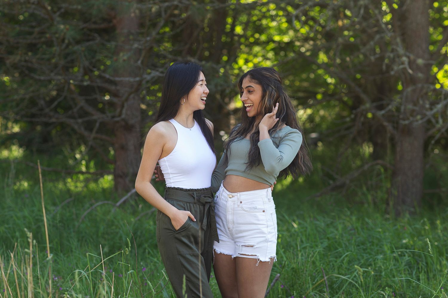 Two young women smiling and talking together outdoors in a lush green forest setting on a sunny day.