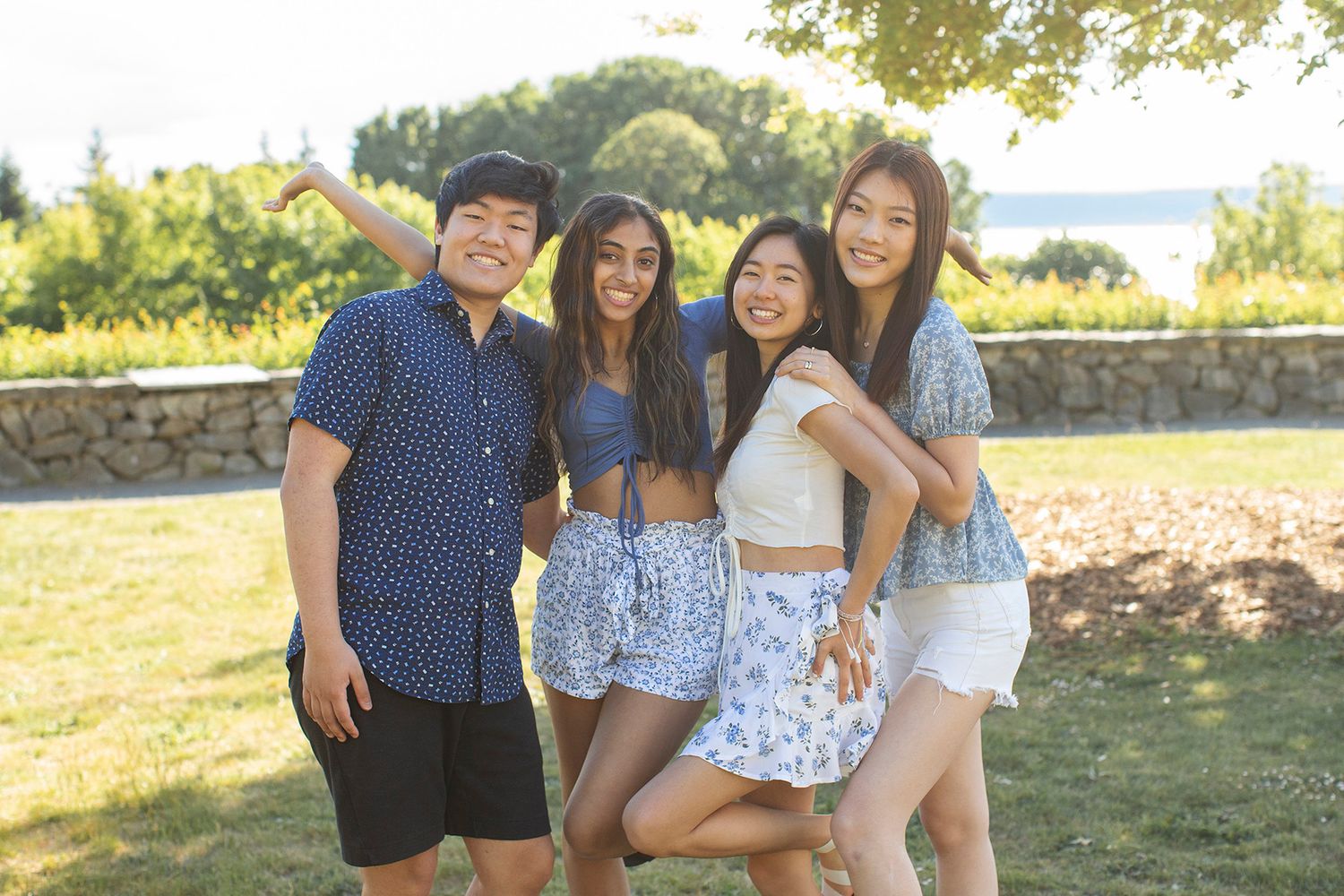 Four smiling teens pose together outdoors in summer outfits, with green trees and a stone wall in the background.