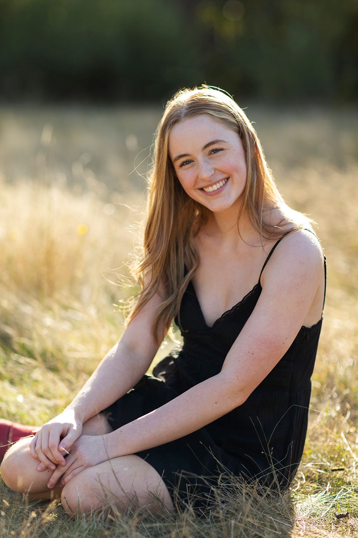 Young woman with long blonde hair smiling in a black dress, sitting cross-legged in a sunlit golden field.