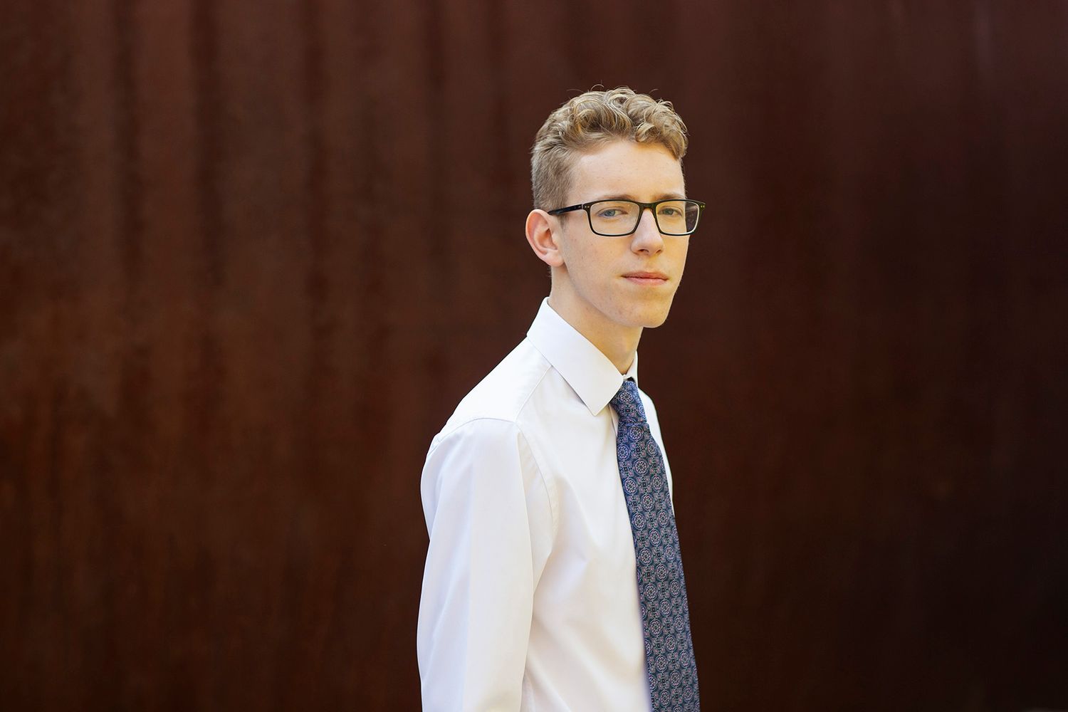 Young man in white shirt and tie with glasses posing outdoors against a rustic brown metal wall.