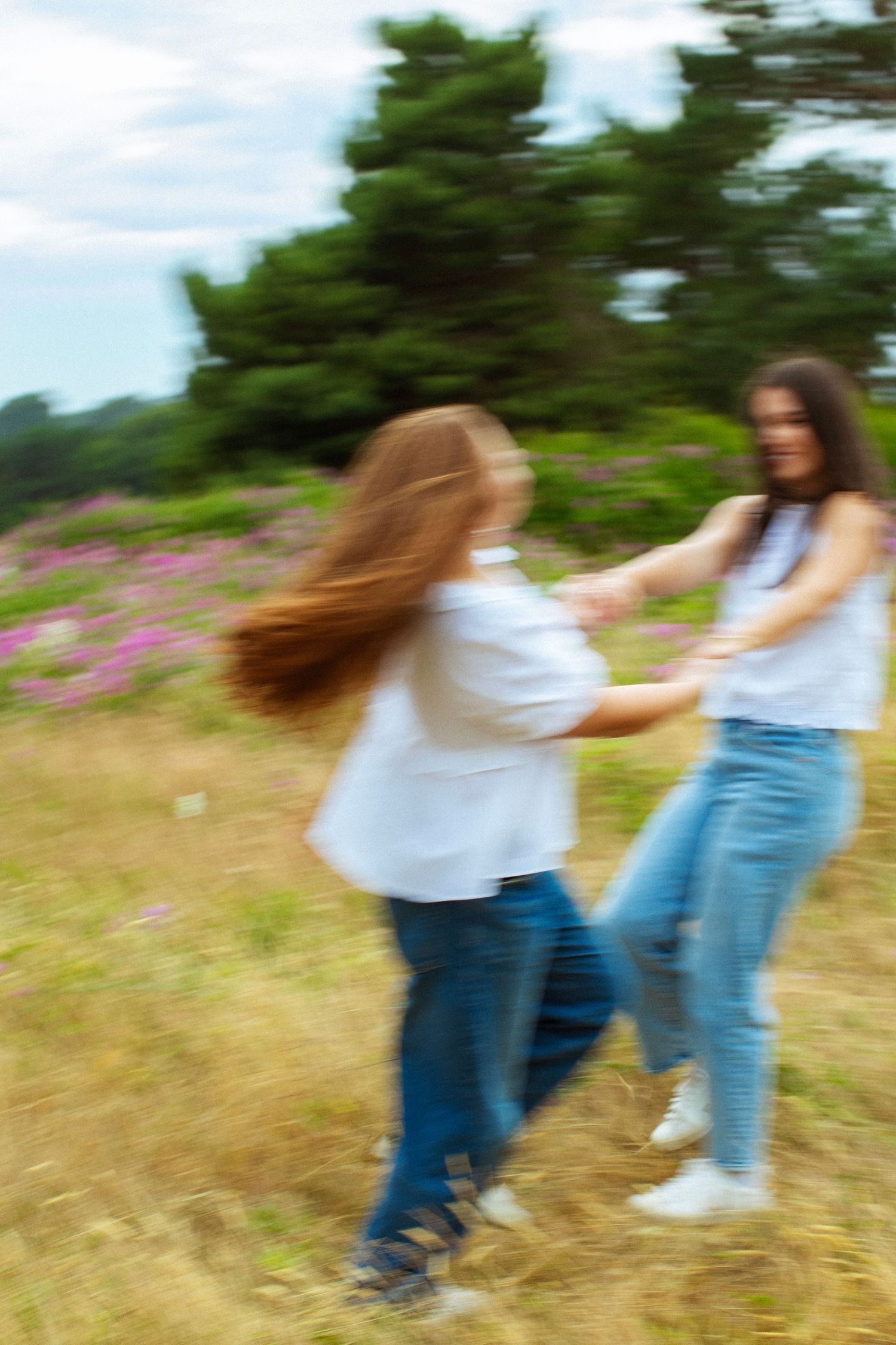 Two girls in white shirts and jeans spin and dance together in a field with pink wildflowers and green trees.