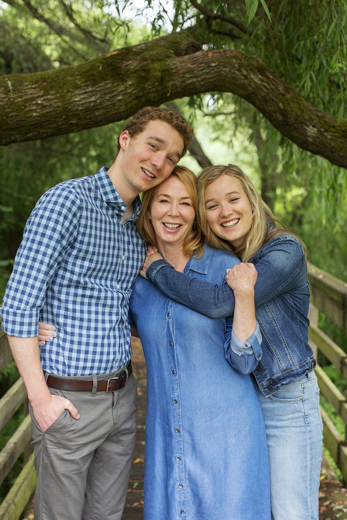 Happy family of three smiling and embracing on a wooden bridge under a weeping willow tree outdoors.