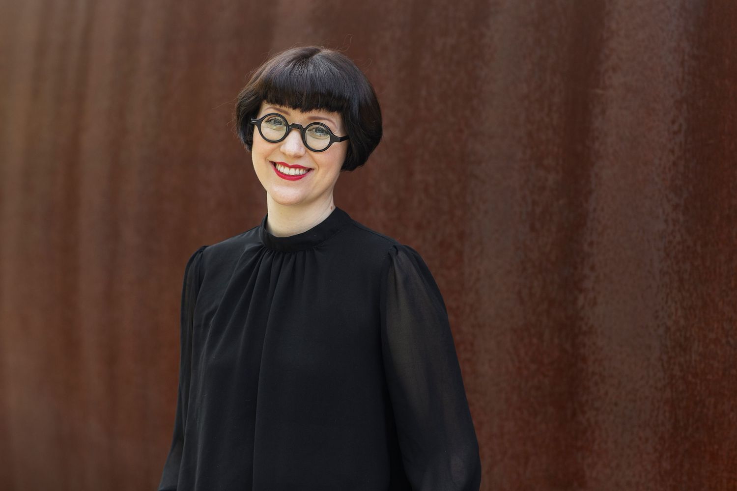 Stylish woman with short dark hair and glasses wearing black dress outdoors.