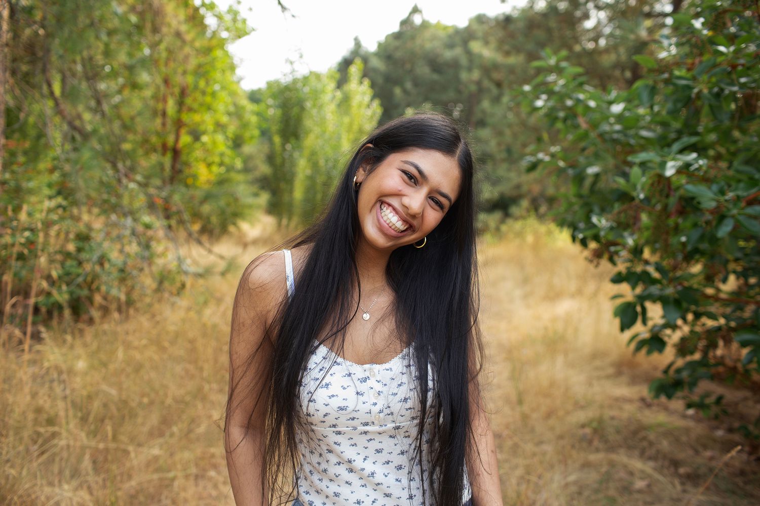 Cheerful young woman with long dark hair smiling on a sunlit woodland trail in summer.