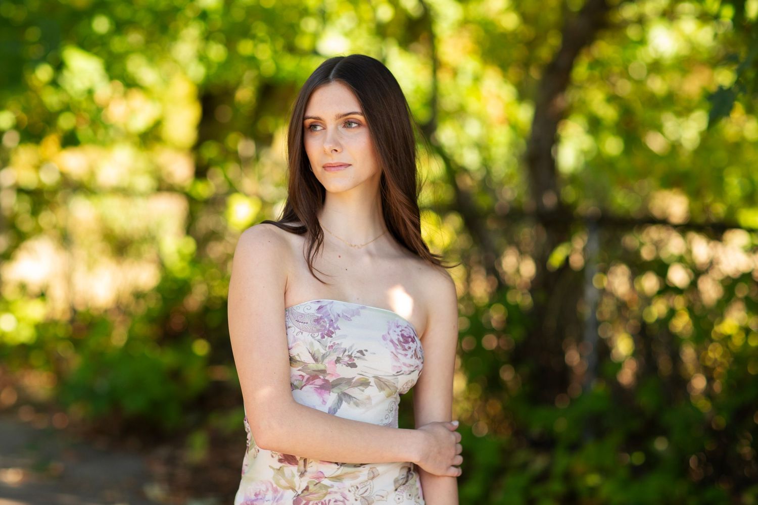 Young woman in floral strapless dress poses outdoors among lush green trees in soft natural light.