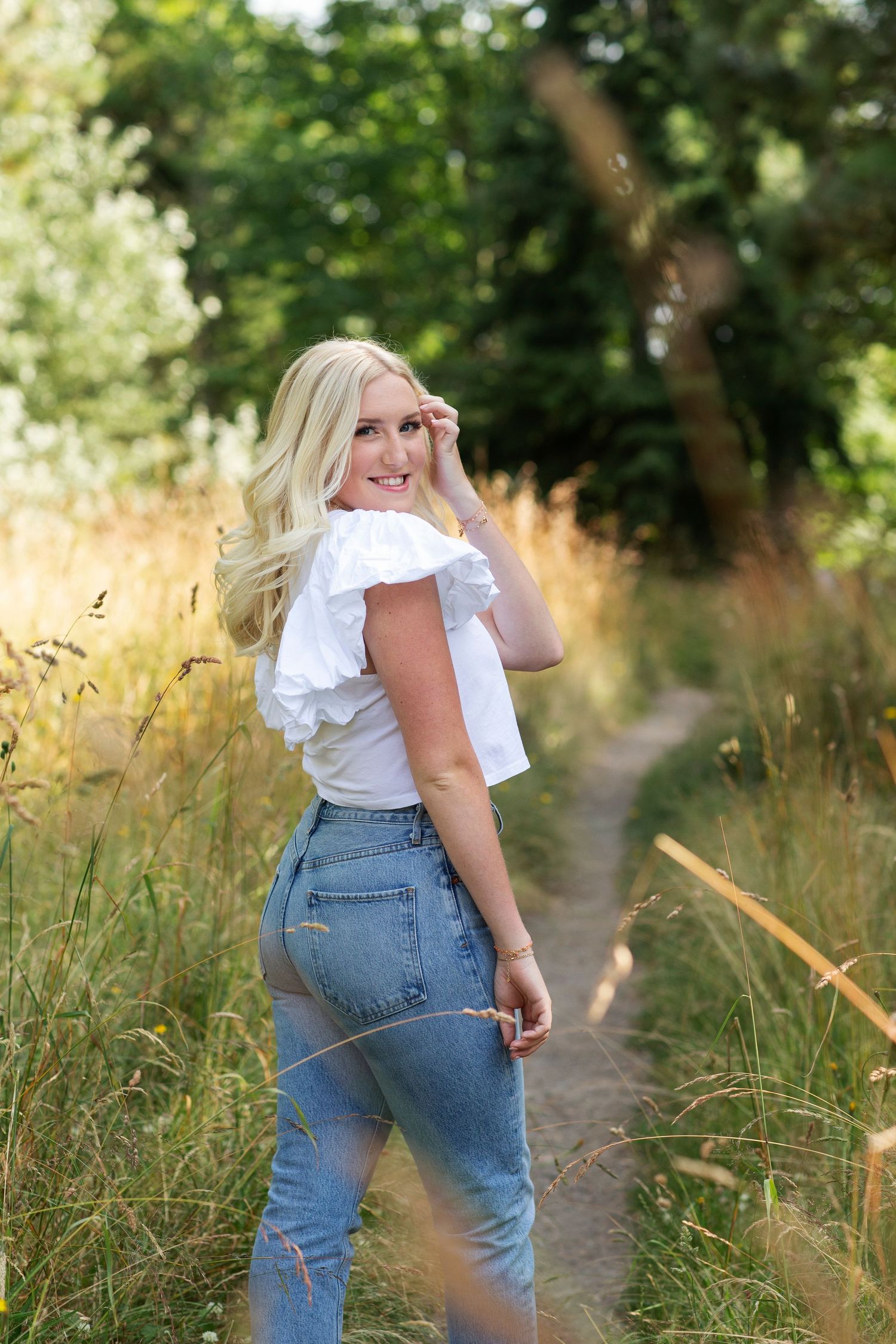 Smiling blonde woman in white ruffle top and jeans posing on a scenic outdoor path with greenery.
