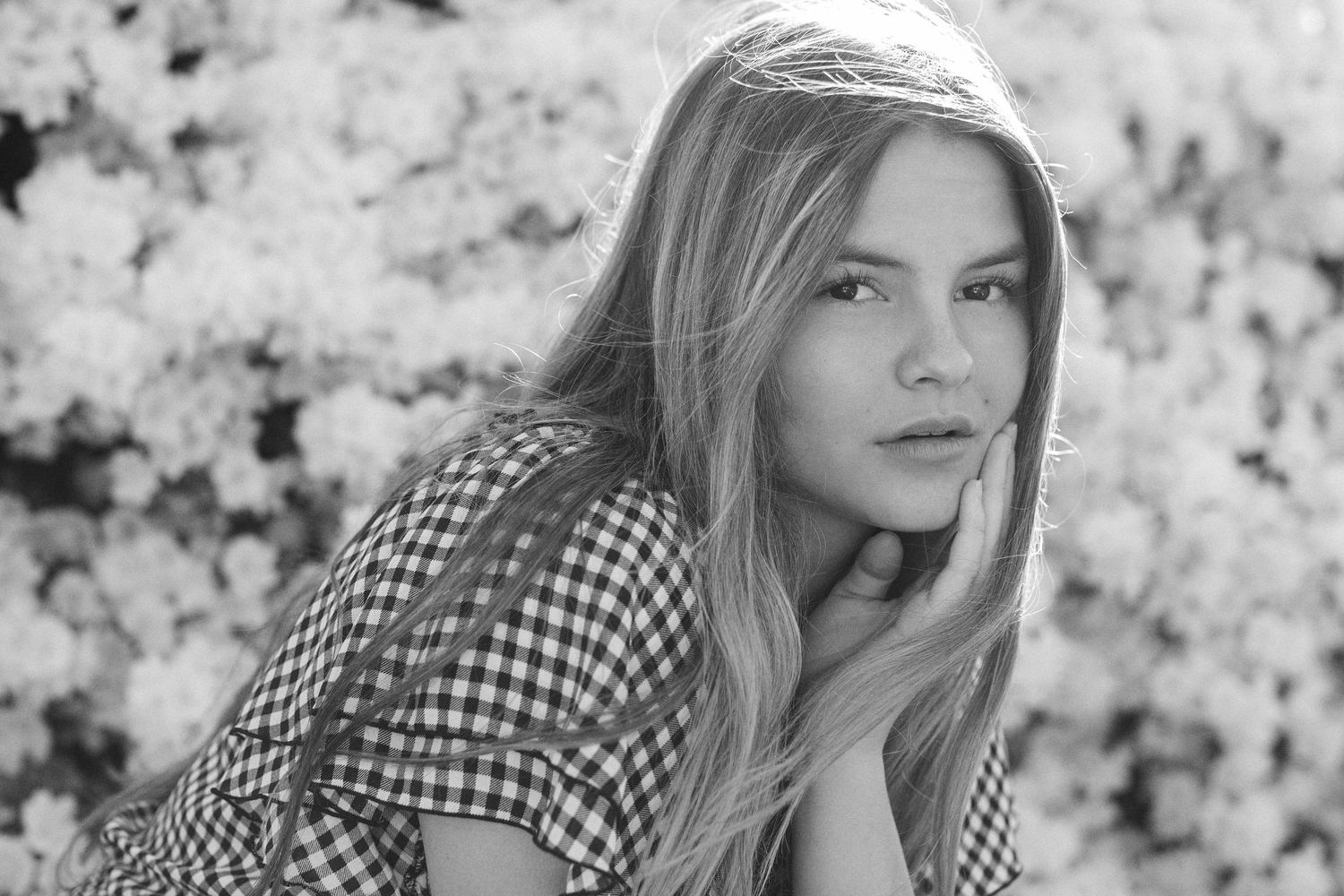 Young woman with long blonde hair in gingham top poses thoughtfully among blooming white flowers.