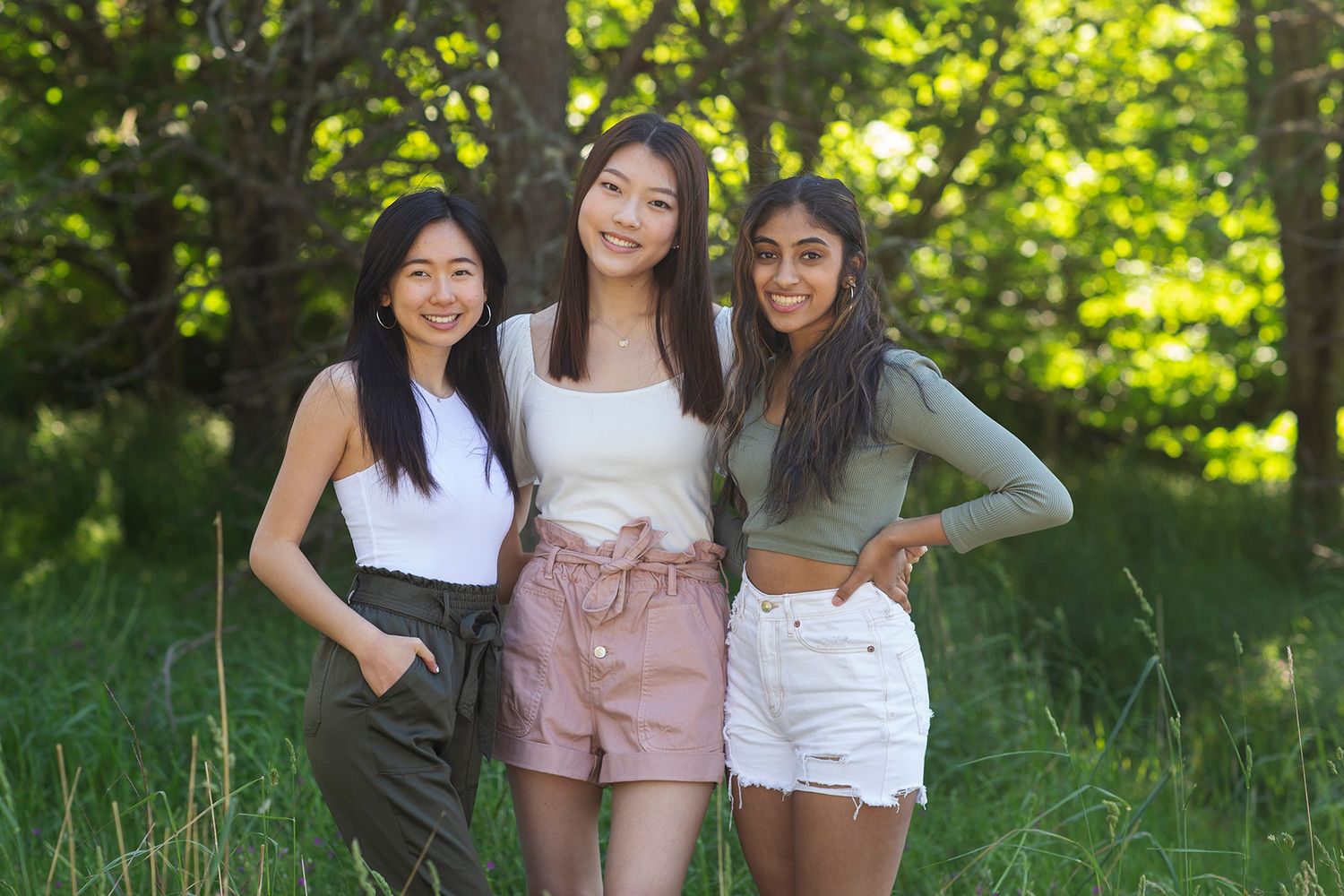 Three young women smiling and posing together outdoors in a lush green park setting on a sunny day.