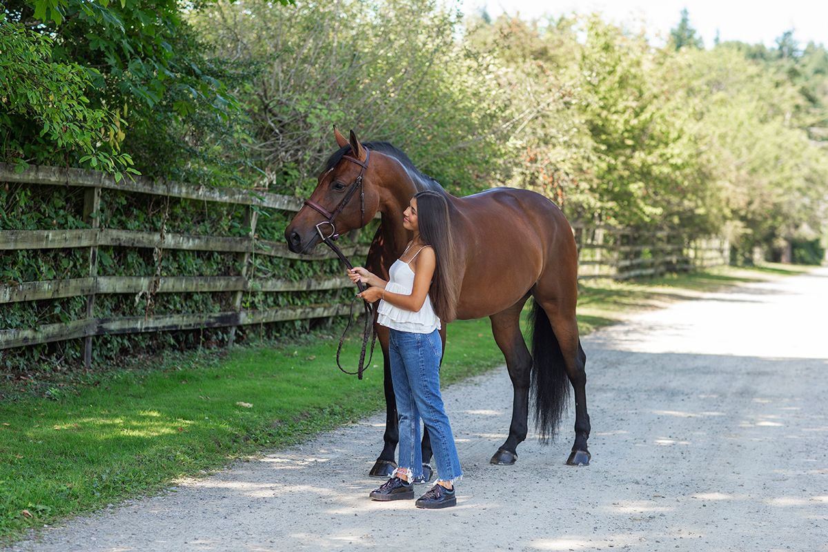 Young girl standing beside a large brown horse on a gravel path near a wooden fence.