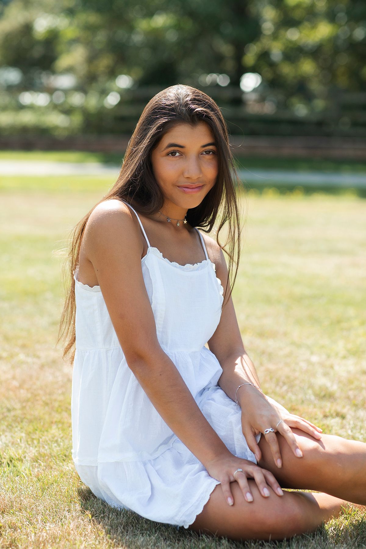 Young woman in white sundress sitting on grass in a sunny park, smiling at camera with long dark hair.