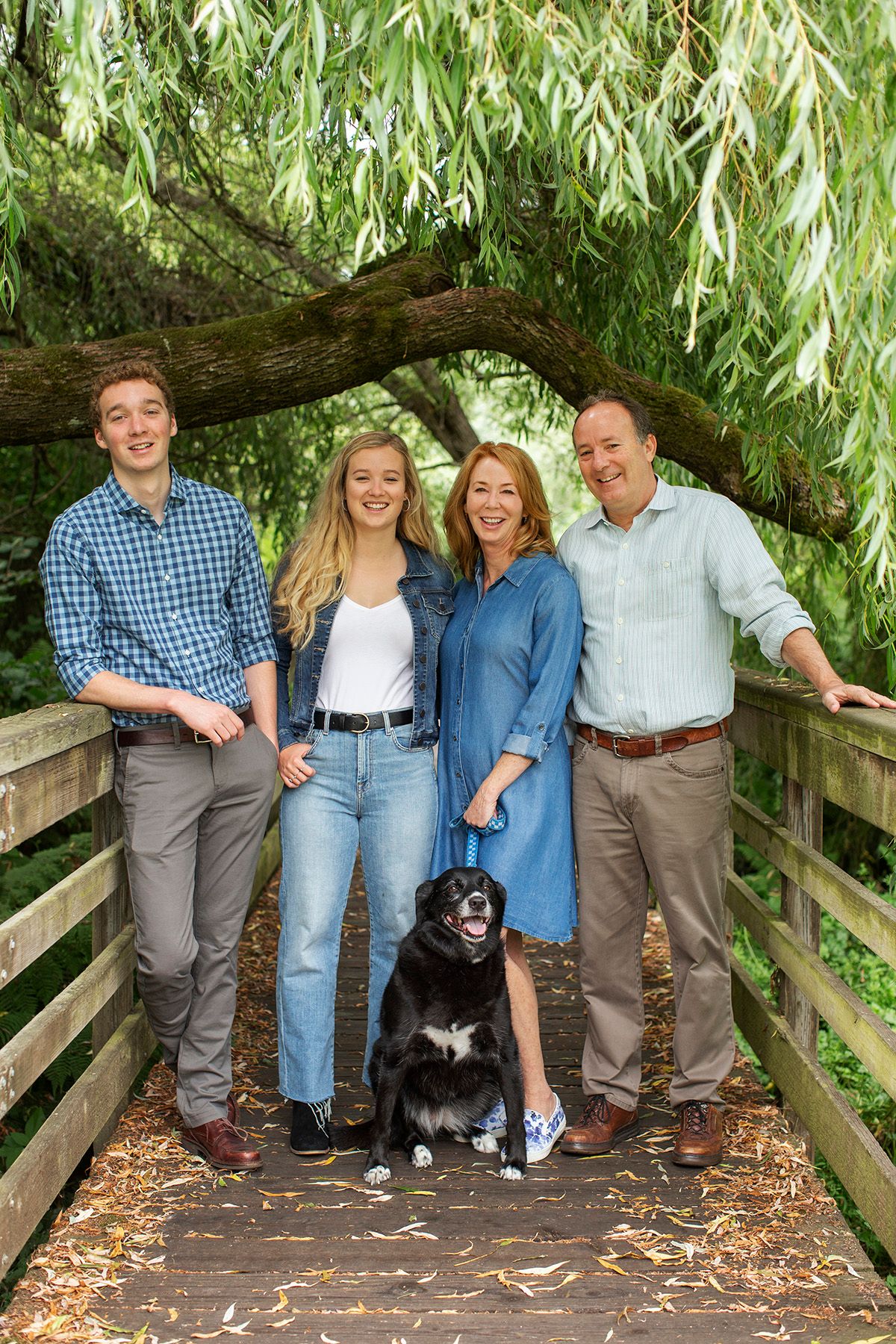 Family of four smiling on a wooden bridge with their black dog, surrounded by lush green trees.
