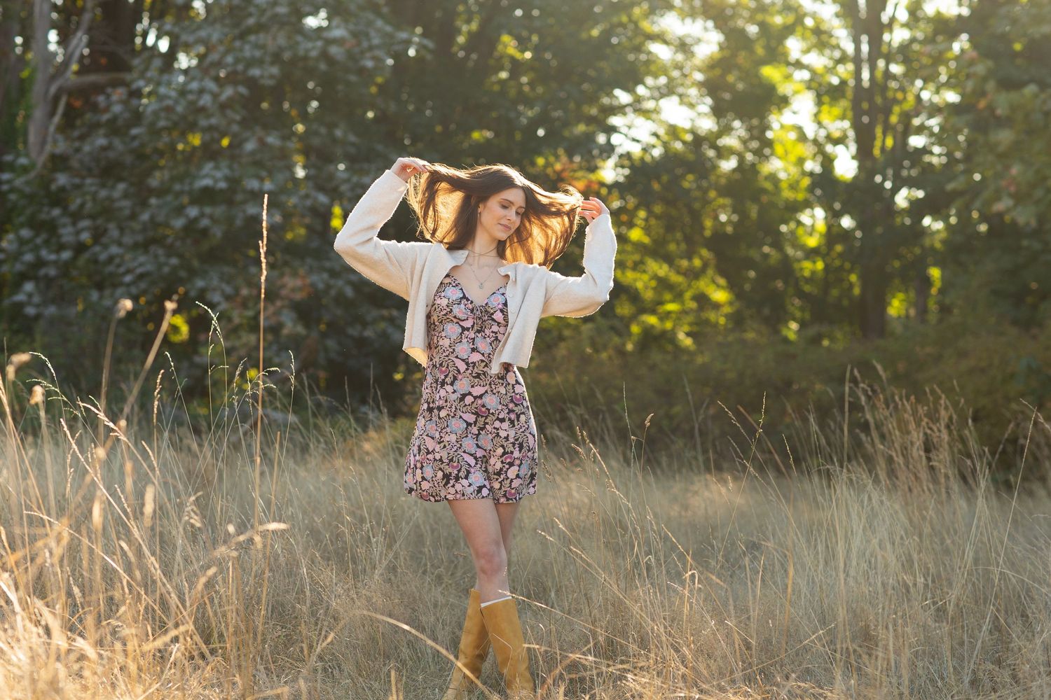 Young woman in floral dress and white cardigan plays with hair in sunlit golden grass field.