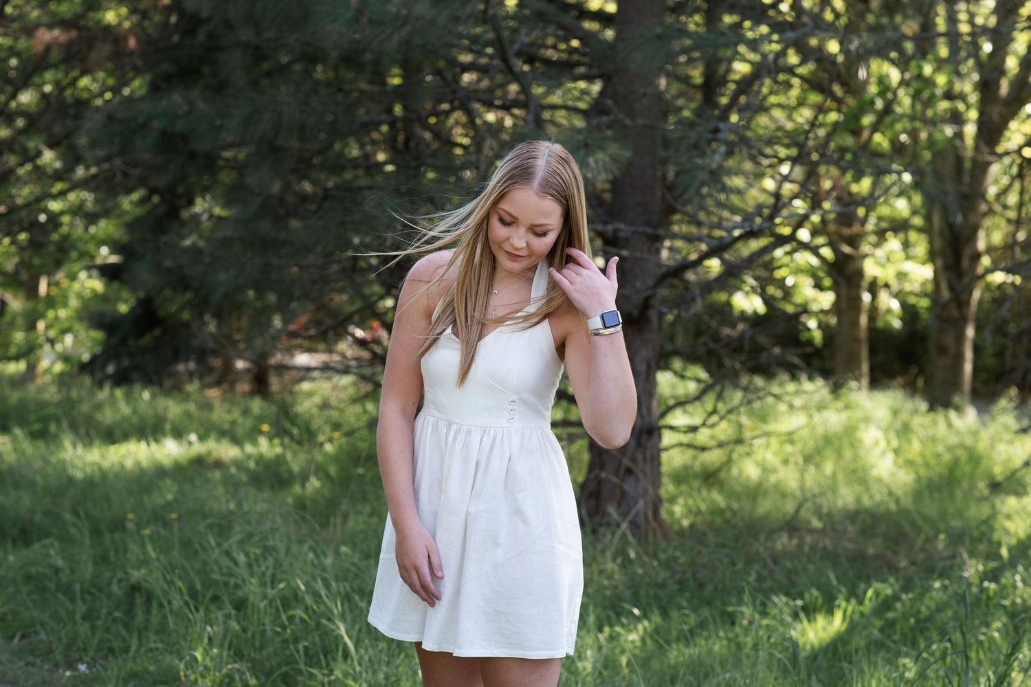 Young woman in white sundress smiling outdoors in green forest setting on a sunny day.