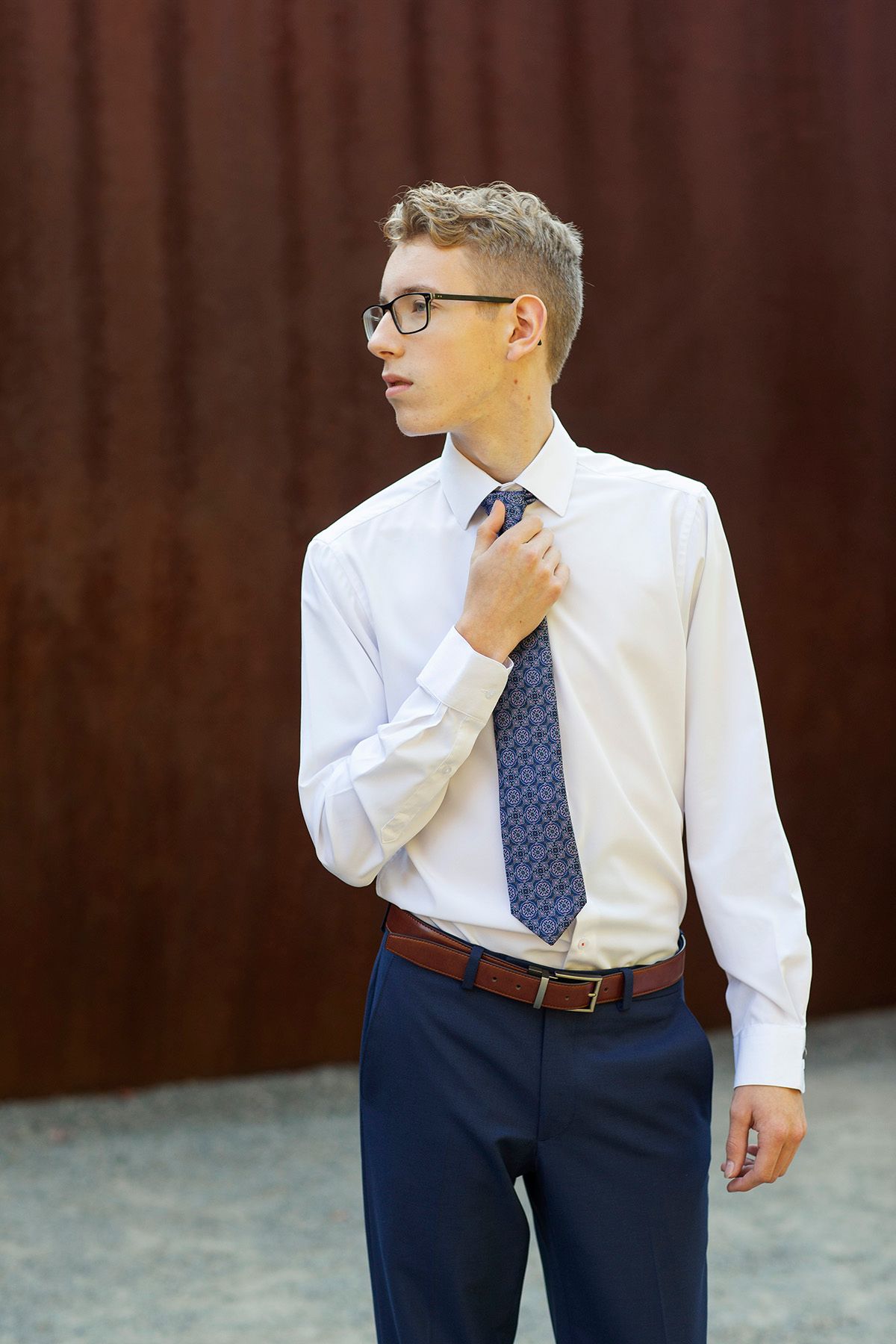 Young businessman in white shirt, blue tie, and glasses adjusting his tie outdoors near a rustic wall.