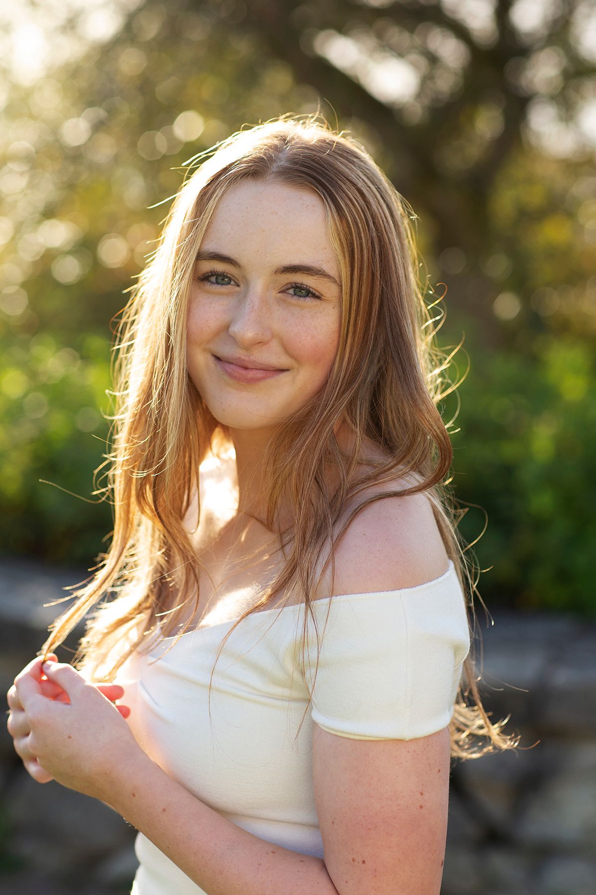 Young woman with long blonde hair smiling outdoors in golden sunlight, wearing an off-shoulder cream top.