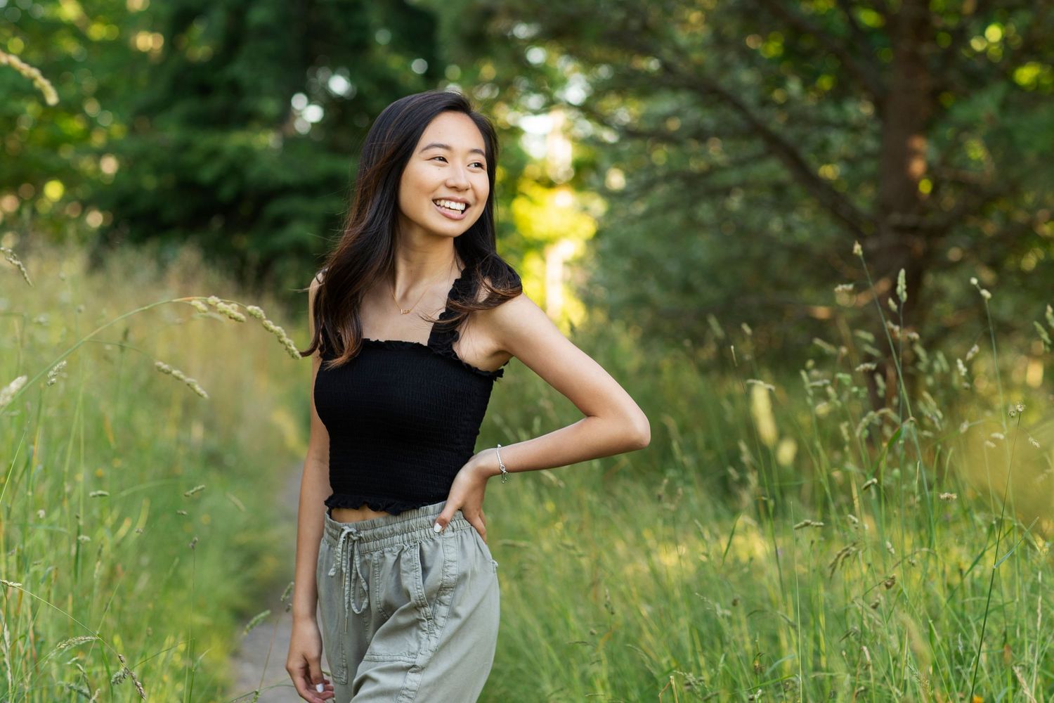 Smiling young Asian woman in black top and gray shorts standing in lush green field with trees in background.