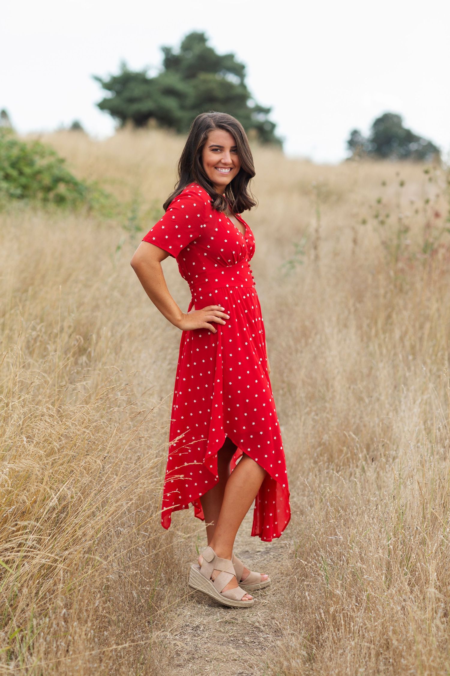 Woman in red polka dot dress standing in golden field, smiling with hand on hip, wearing wedge sandals.