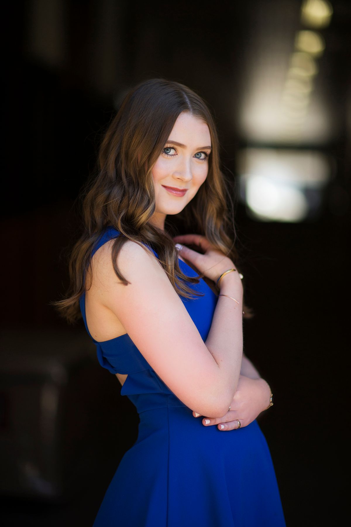 Young woman in royal blue sleeveless dress posing confidently with arms crossed, smiling over shoulder.