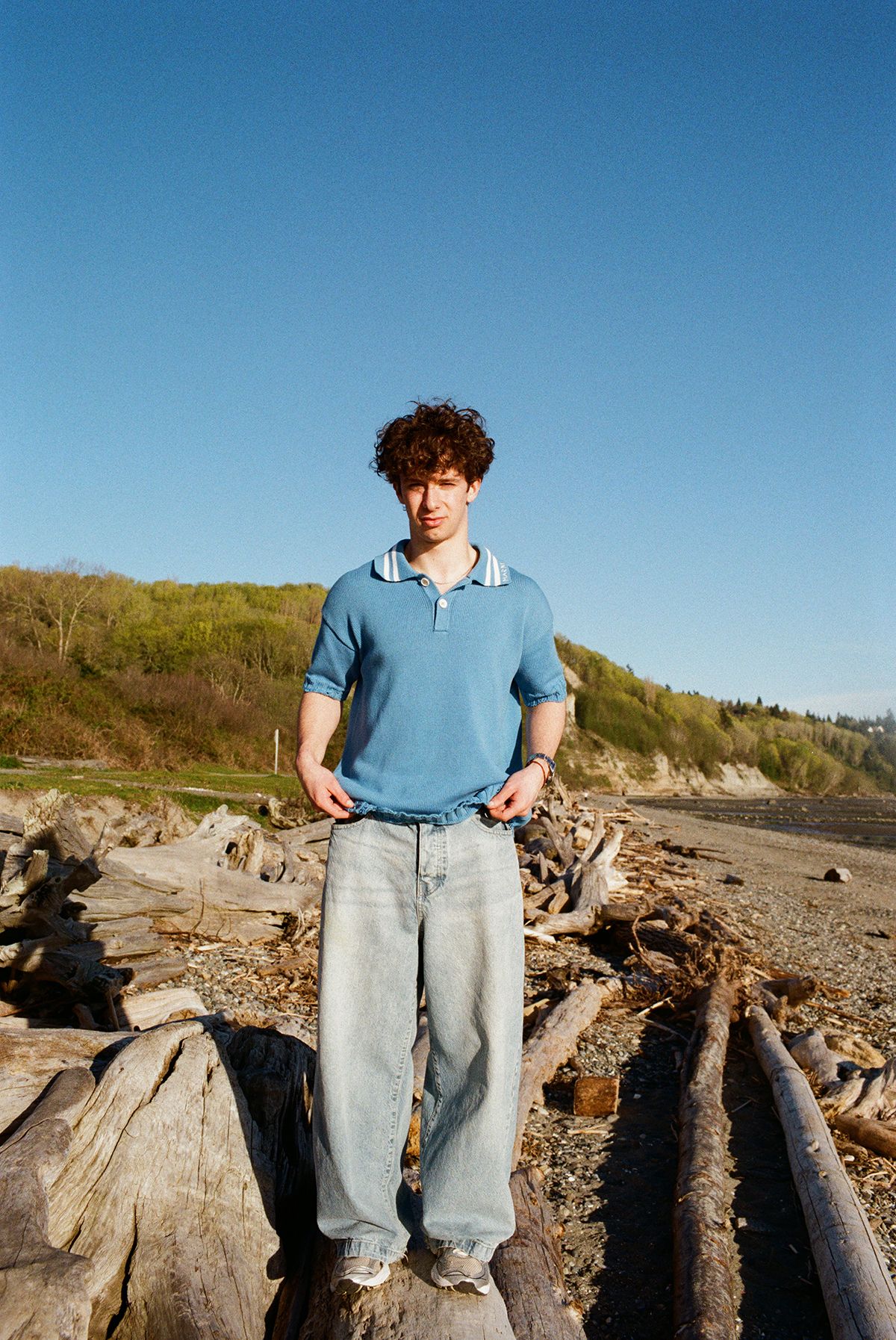 Young man with curly hair in blue polo shirt and jeans standing on driftwood logs at a rocky beach.