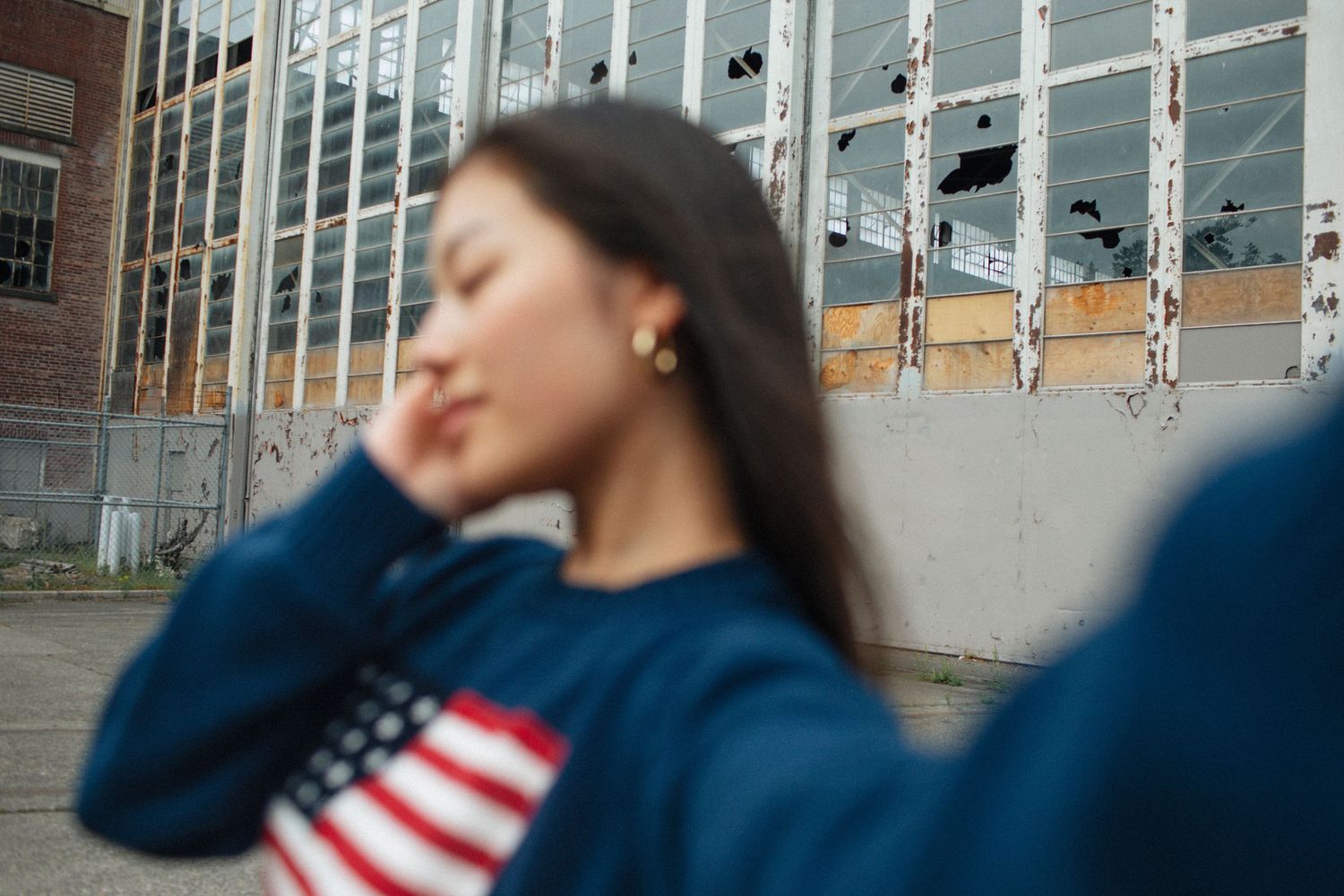 Close-up of young woman in American flag sweater with eyes closed near vintage industrial building.