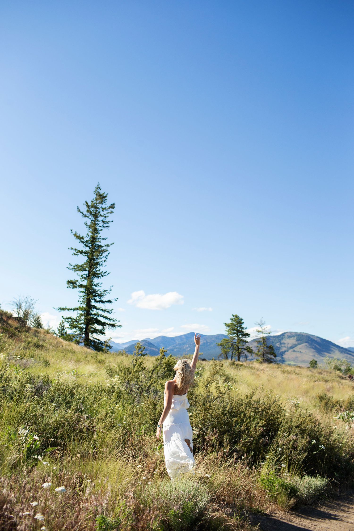 Blonde woman in white dress reaching up toward the sky near a tall pine tree in mountains.