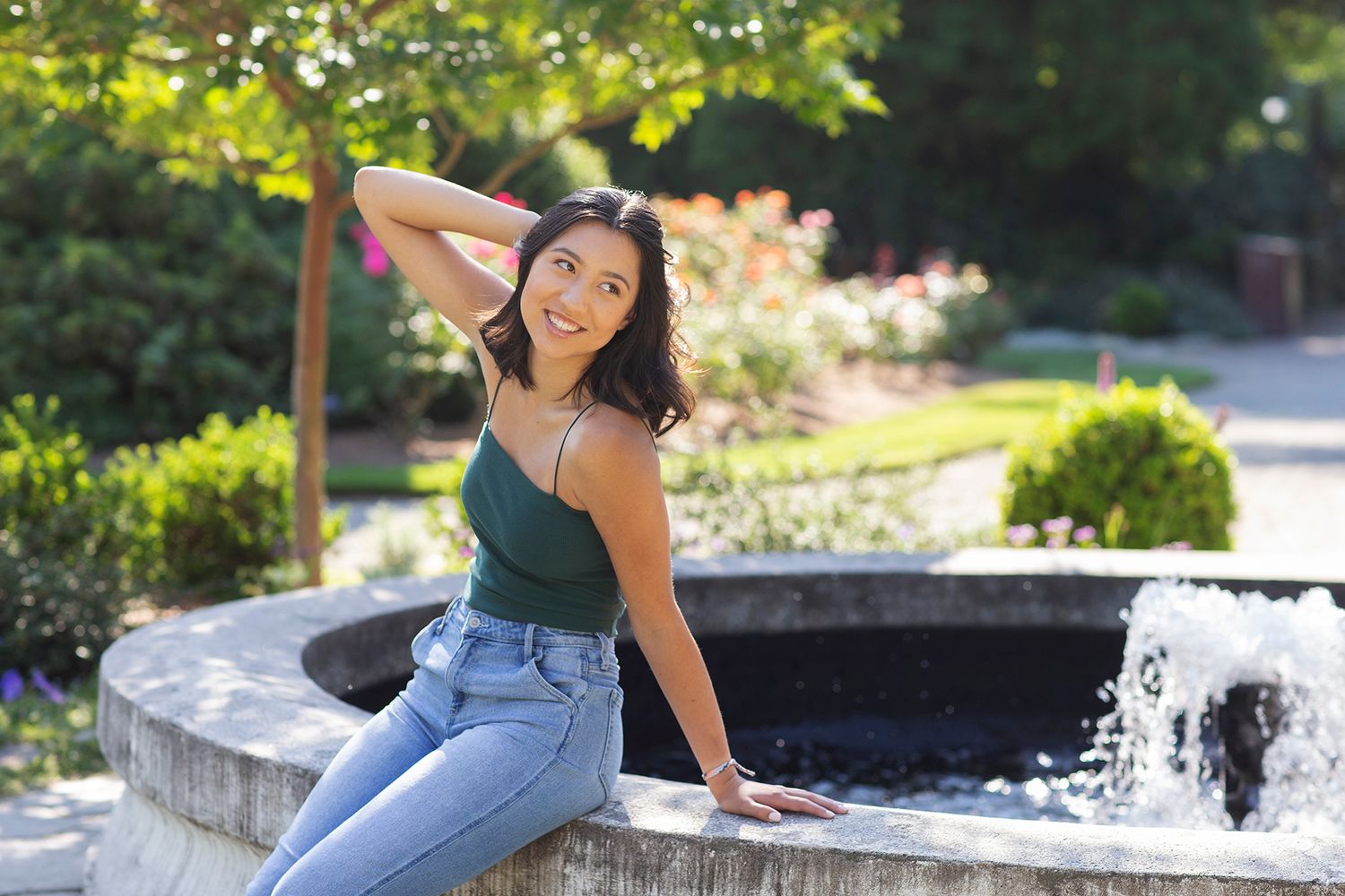 Young Asian woman in green tank top and jeans sits by a garden fountain on a sunny day.