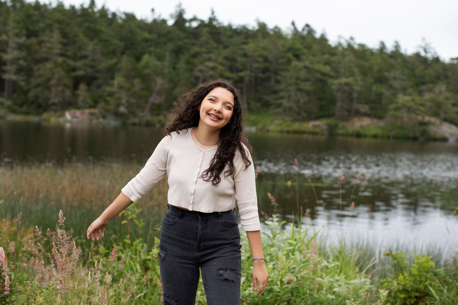 Smiling young woman in cream top standing by a serene lake surrounded by lush greenery.