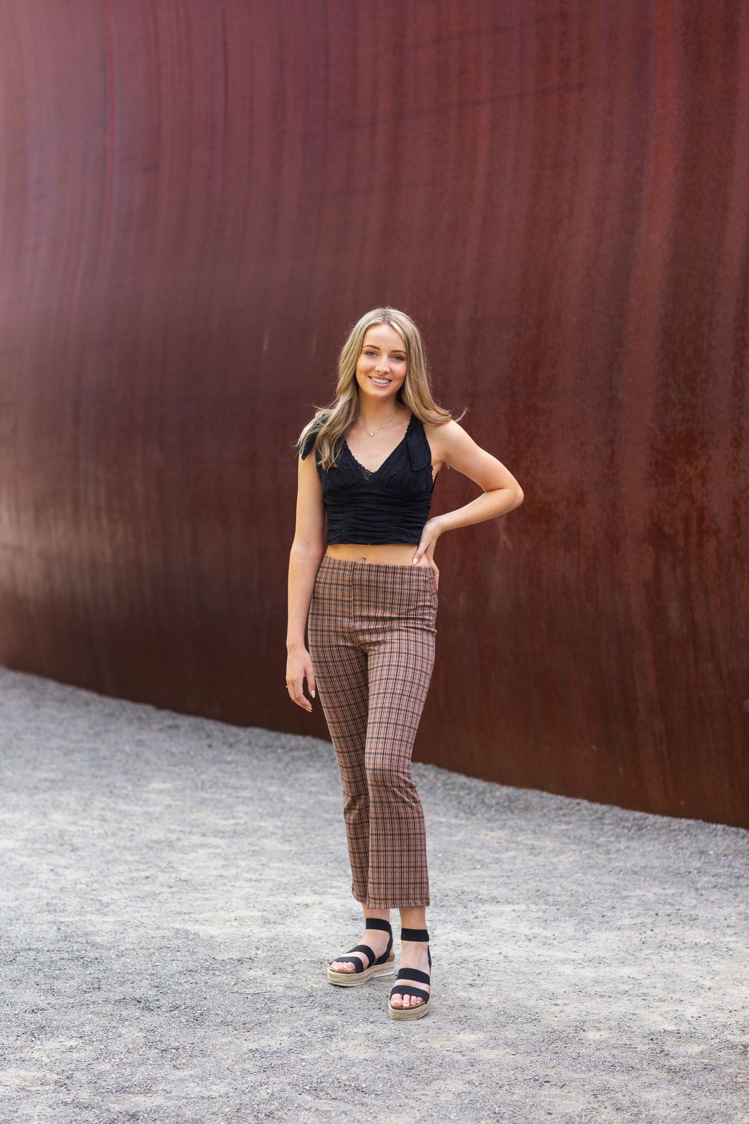 Blonde woman in black crop top and plaid pants smiling against rusty metal wall.