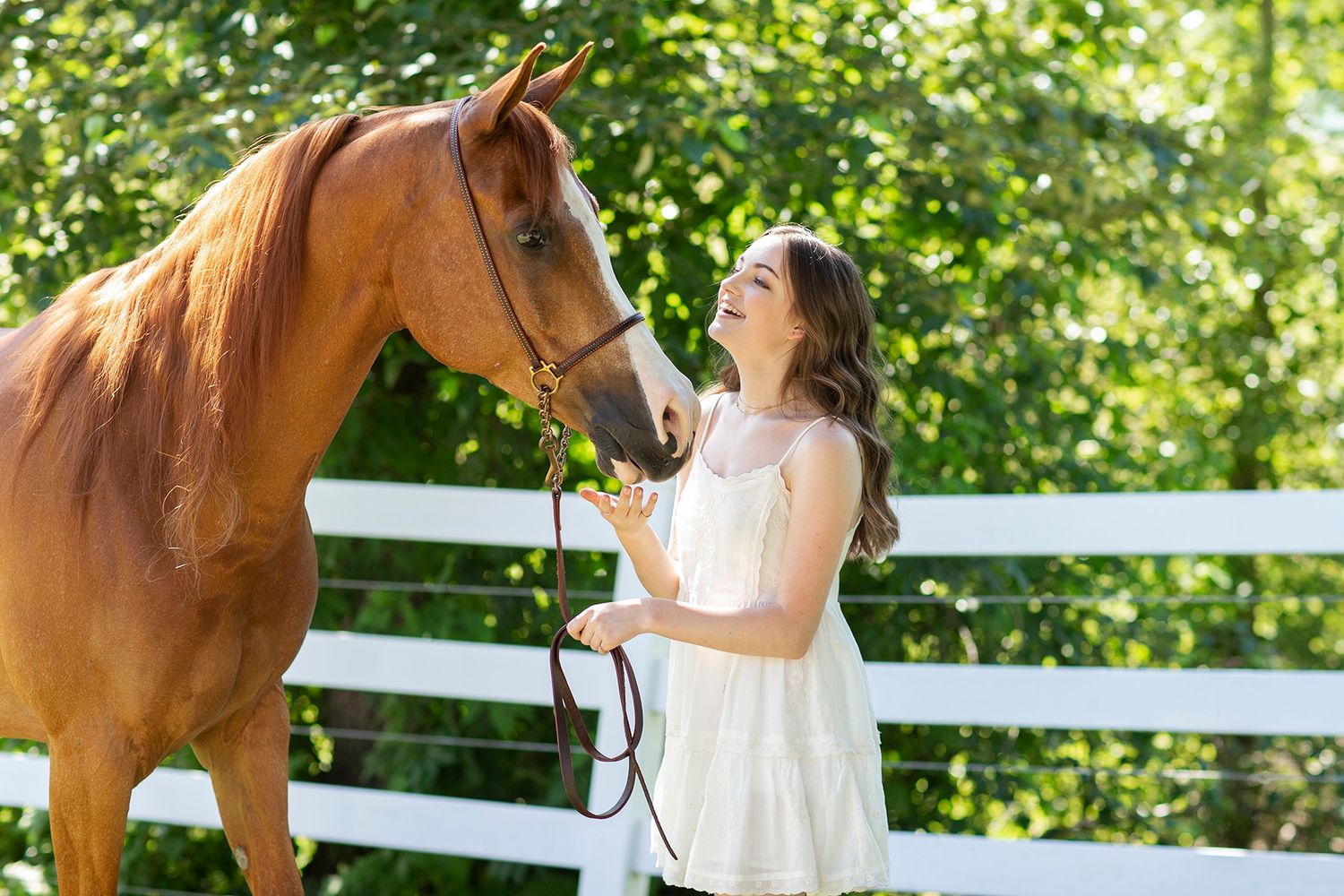 A smiling young woman in a white dress holds the reins of a chestnut horse by a white fence outdoors.