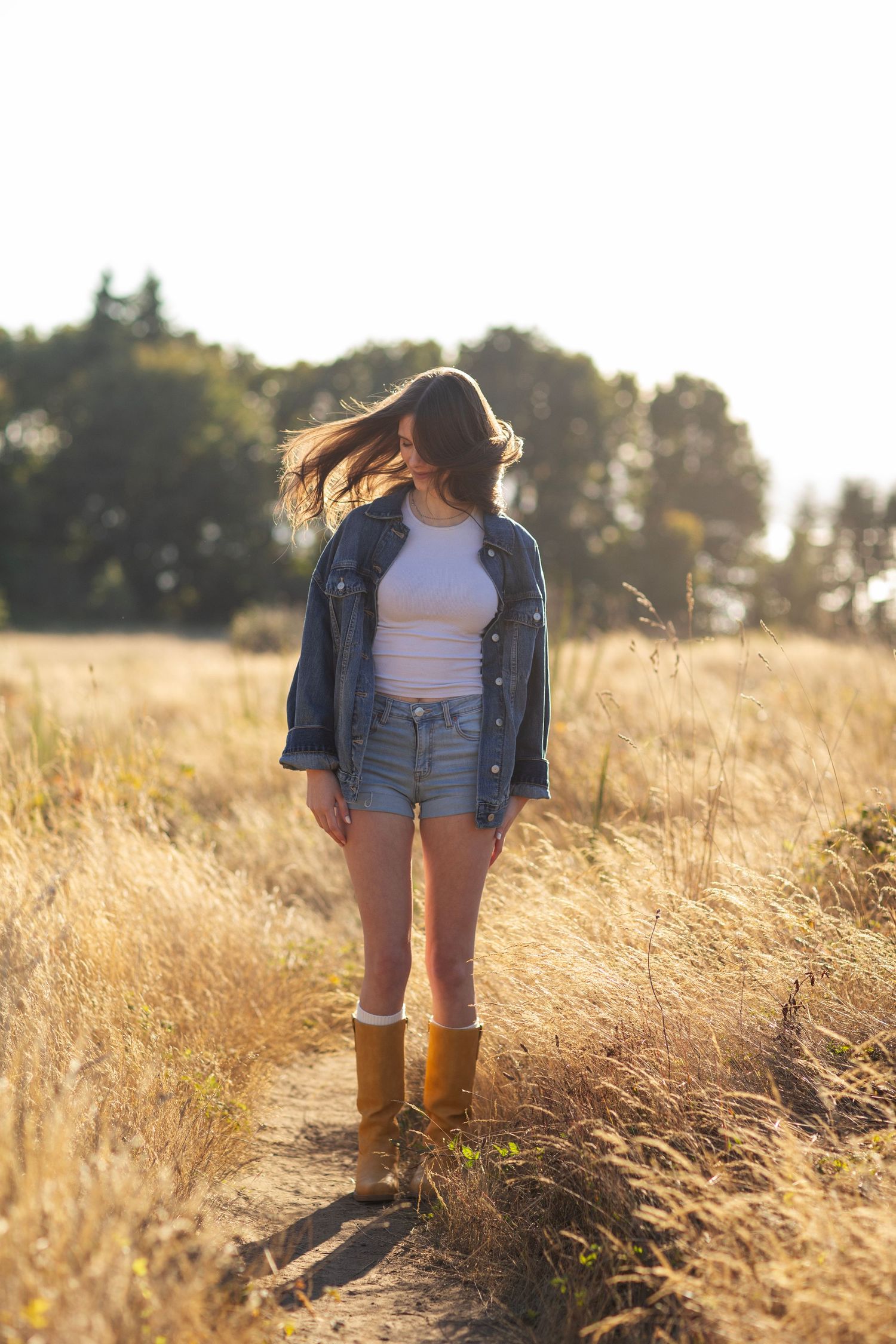 Young woman in denim jacket, shorts, and yellow boots standing on a sunlit field path.
