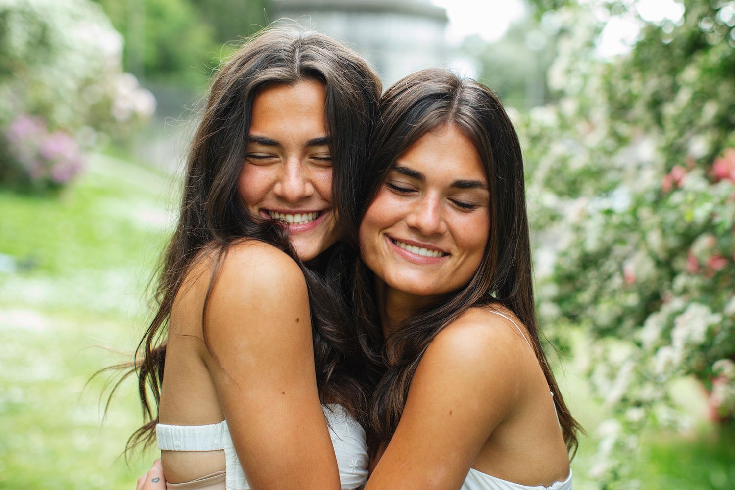 Two young women with long dark hair share a joyful hug outdoors in a lush green garden with blooming flowers.