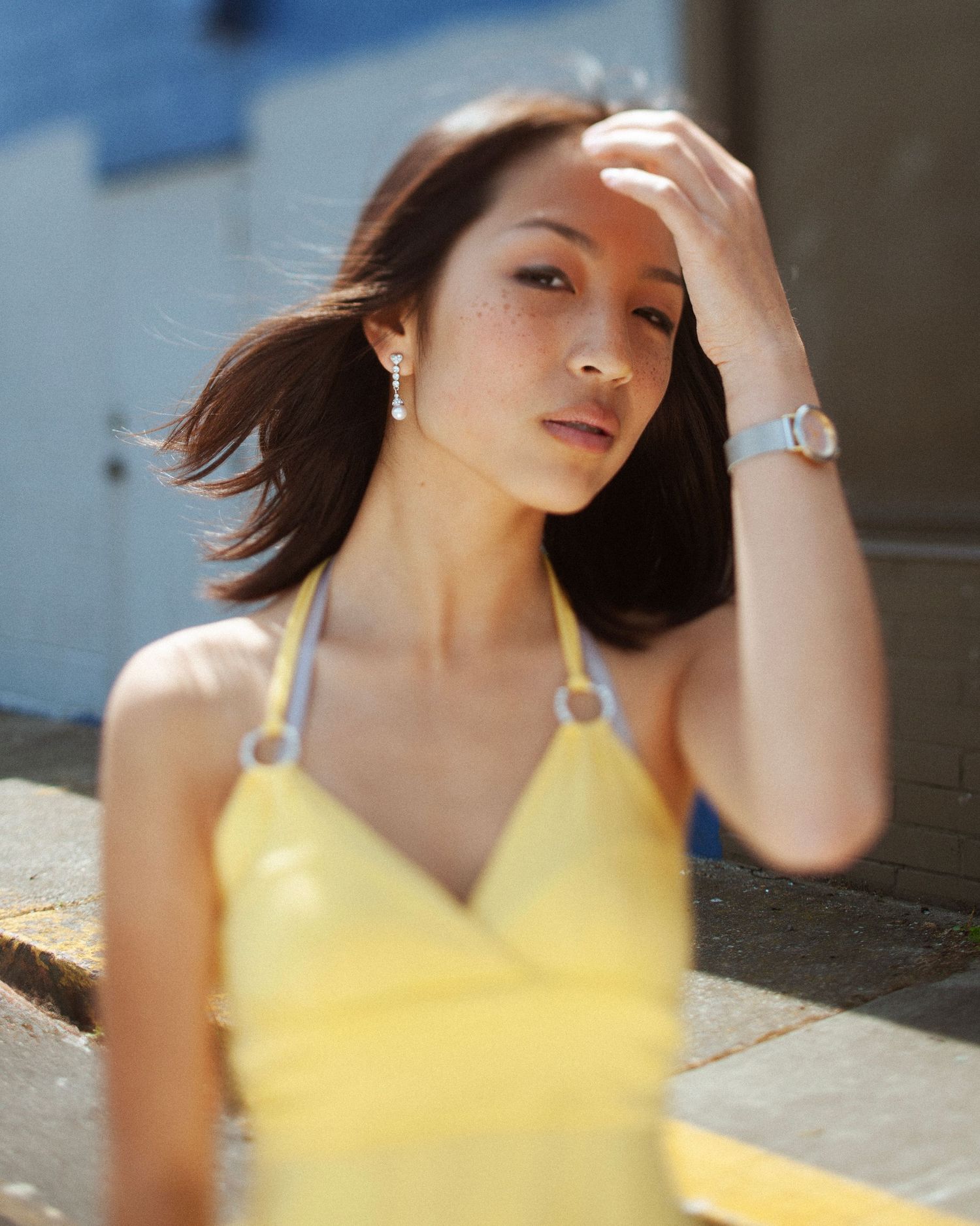Young Asian woman in yellow halter top posing outdoors in sunlight, wearing a silver watch and drop earrings.