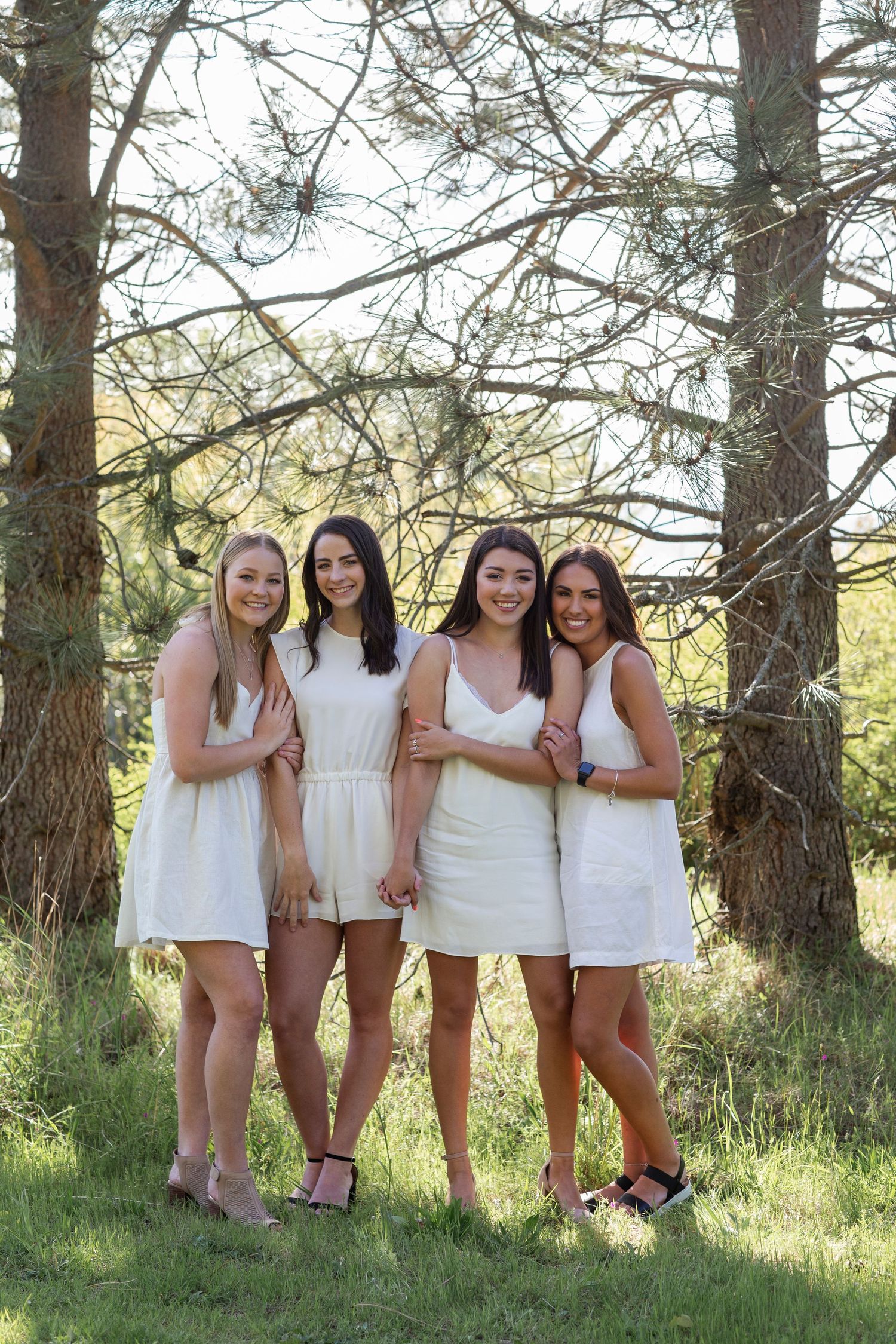 Four young women in white dresses pose together outdoors among pine trees on a sunny day.