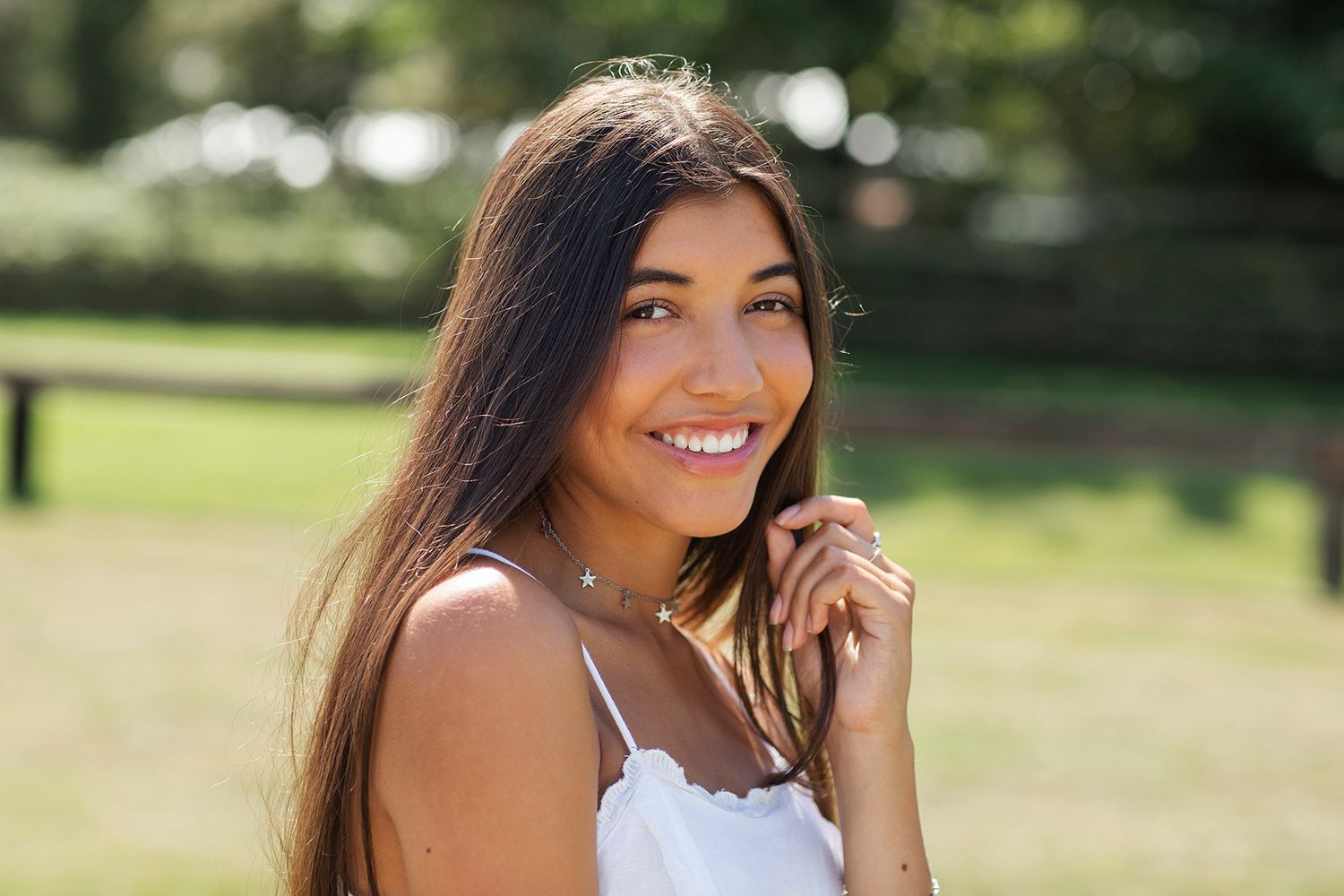 Young woman with long brown hair posing outdoors under green tree leaves in soft natural sunlight.