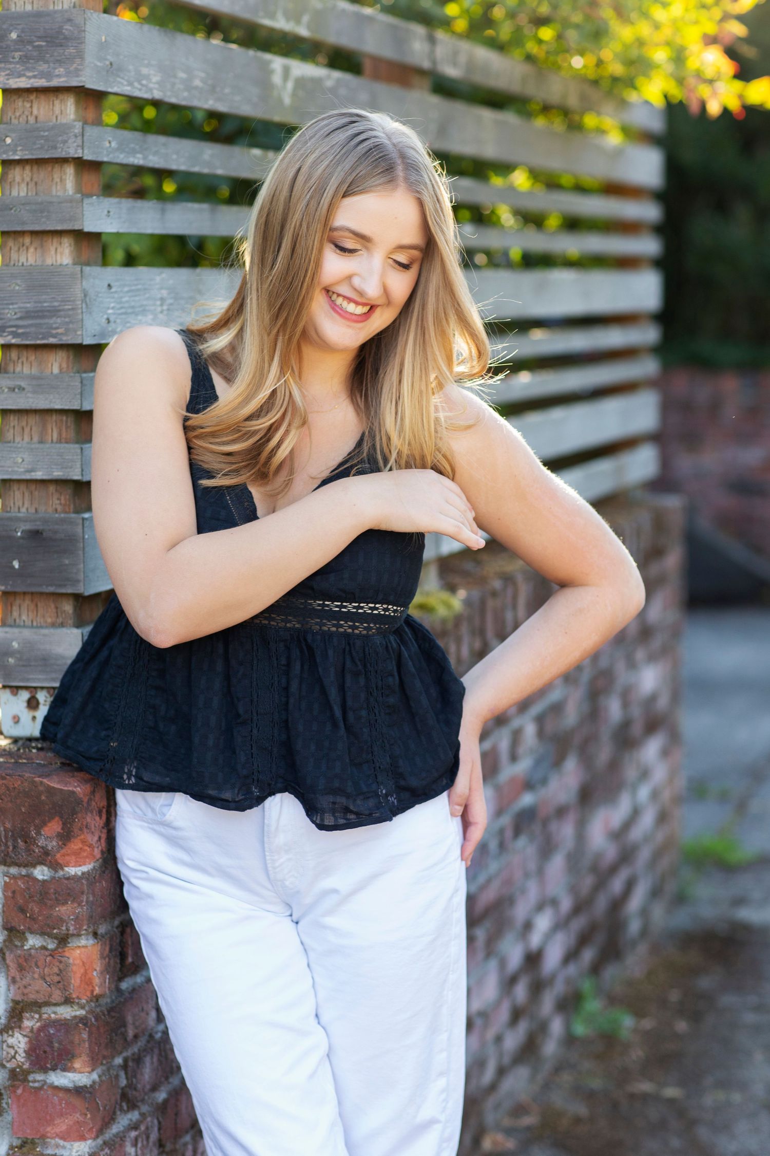 Smiling blonde woman in black top and white pants leaning against a brick and wood fence.