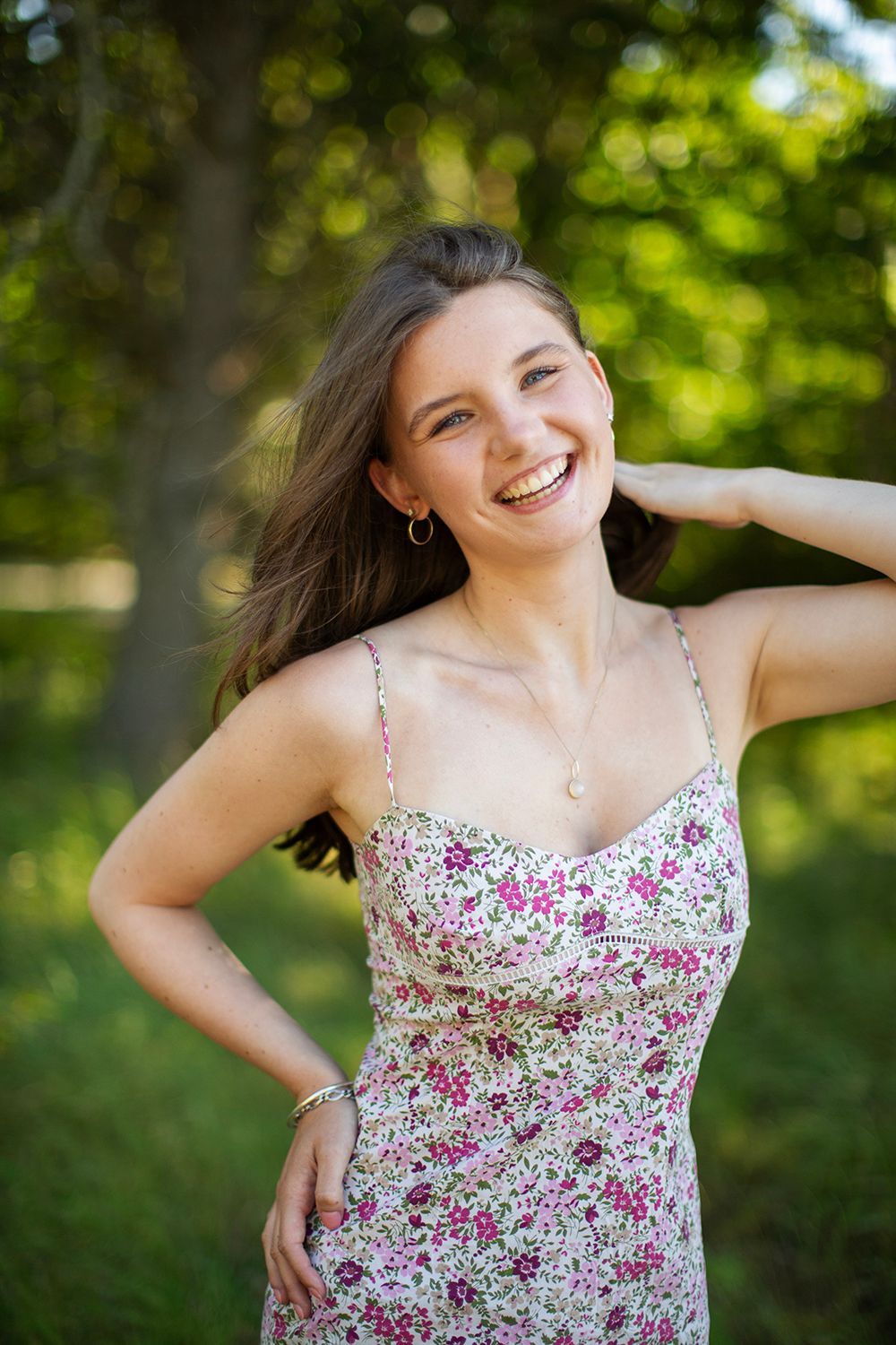 Young woman in floral sundress laughing outdoors in green park setting, long brown hair, wearing gold jewelry.