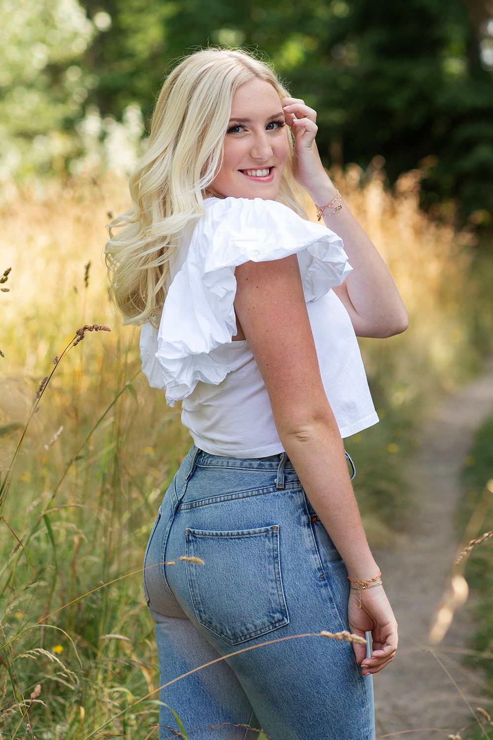 Smiling blonde woman in white ruffle top and jeans poses on a sunny nature trail surrounded by tall golden grasses.