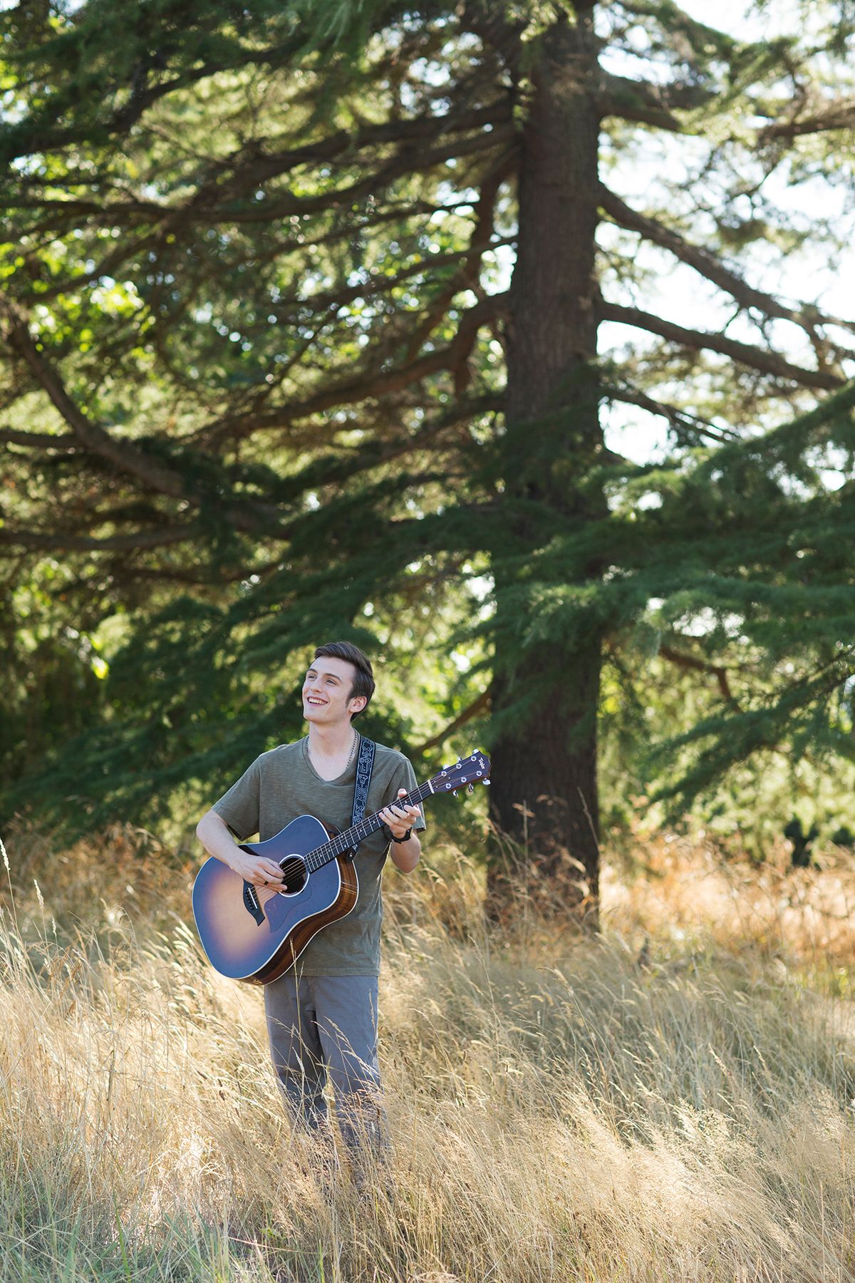 Young man smiling while playing acoustic guitar outdoors in tall grass near a large evergreen tree.
