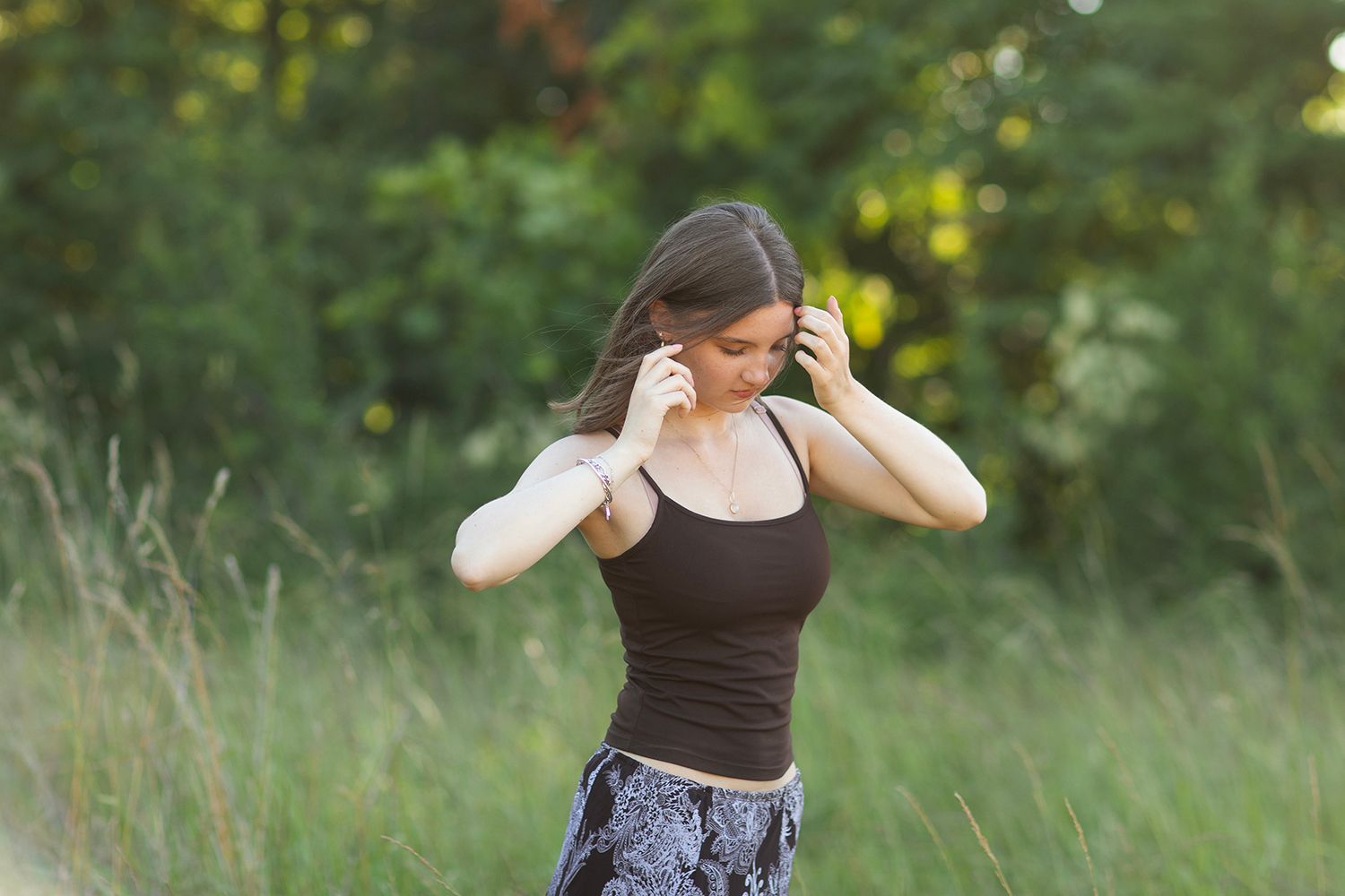 Young woman in black tank top adjusting earphones outdoors in green field with trees in background.