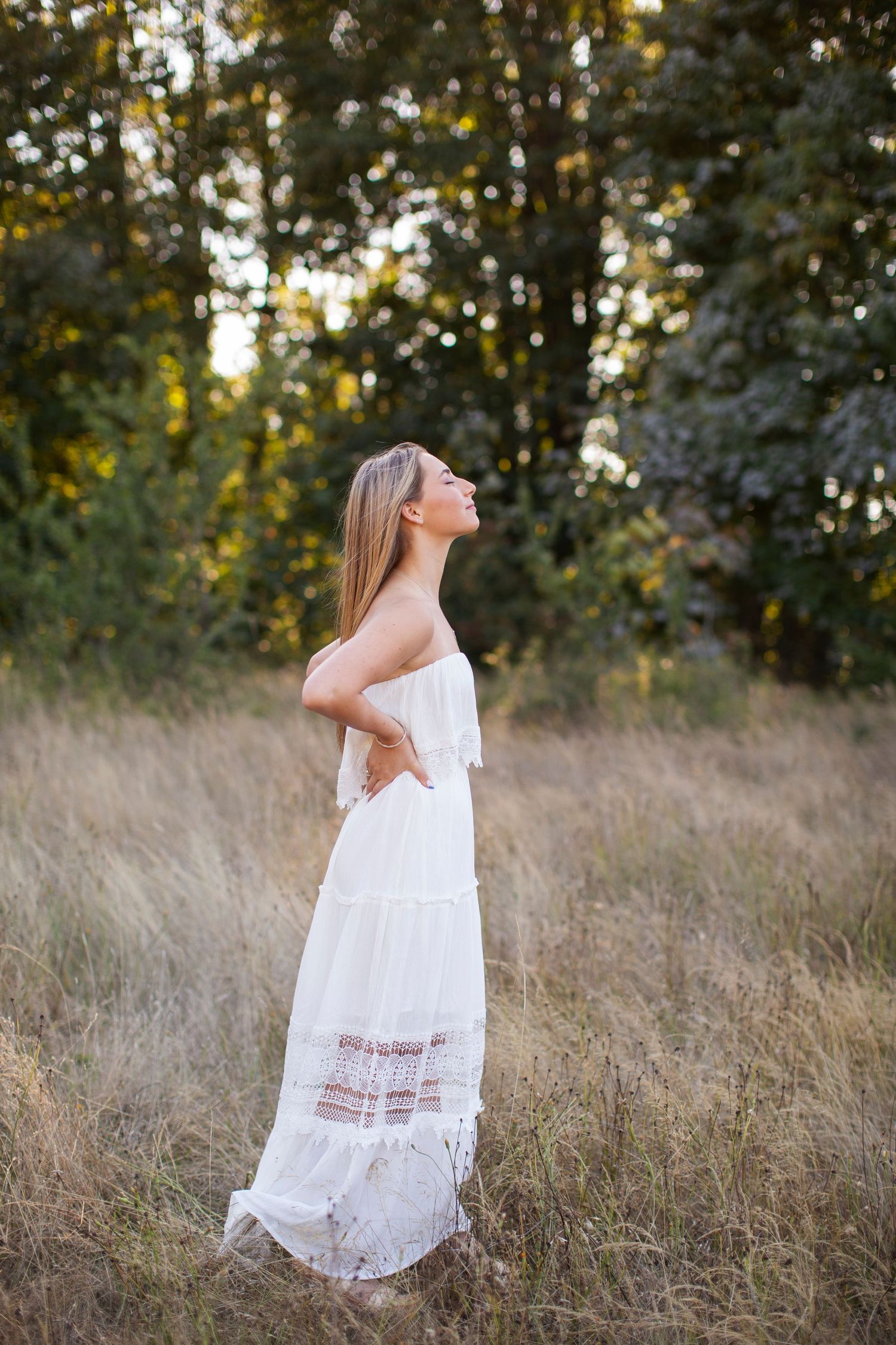 Blonde woman in white strapless tiered maxi dress stands in golden field with forest backdrop at sunset.