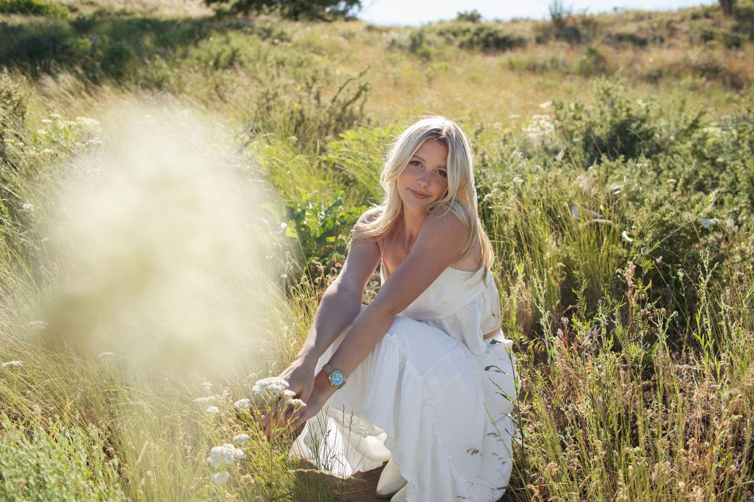 Blonde woman in white dress sitting peacefully in a sunlit meadow with wildflowers.