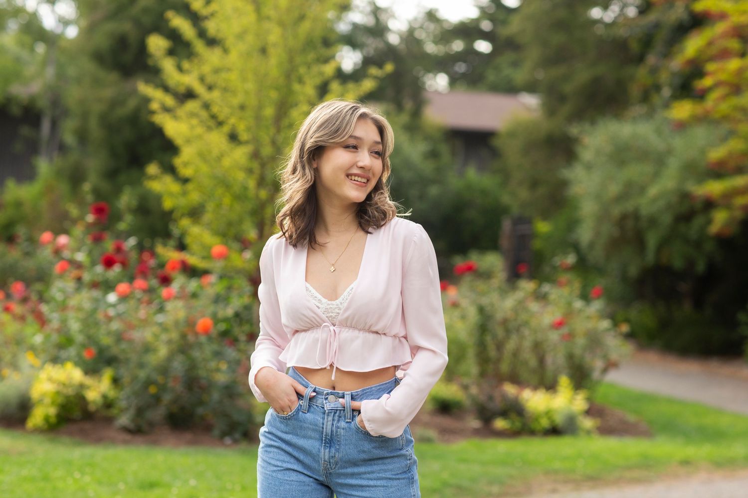 Smiling young woman in pink crop top and jeans standing in a colorful outdoor garden setting.