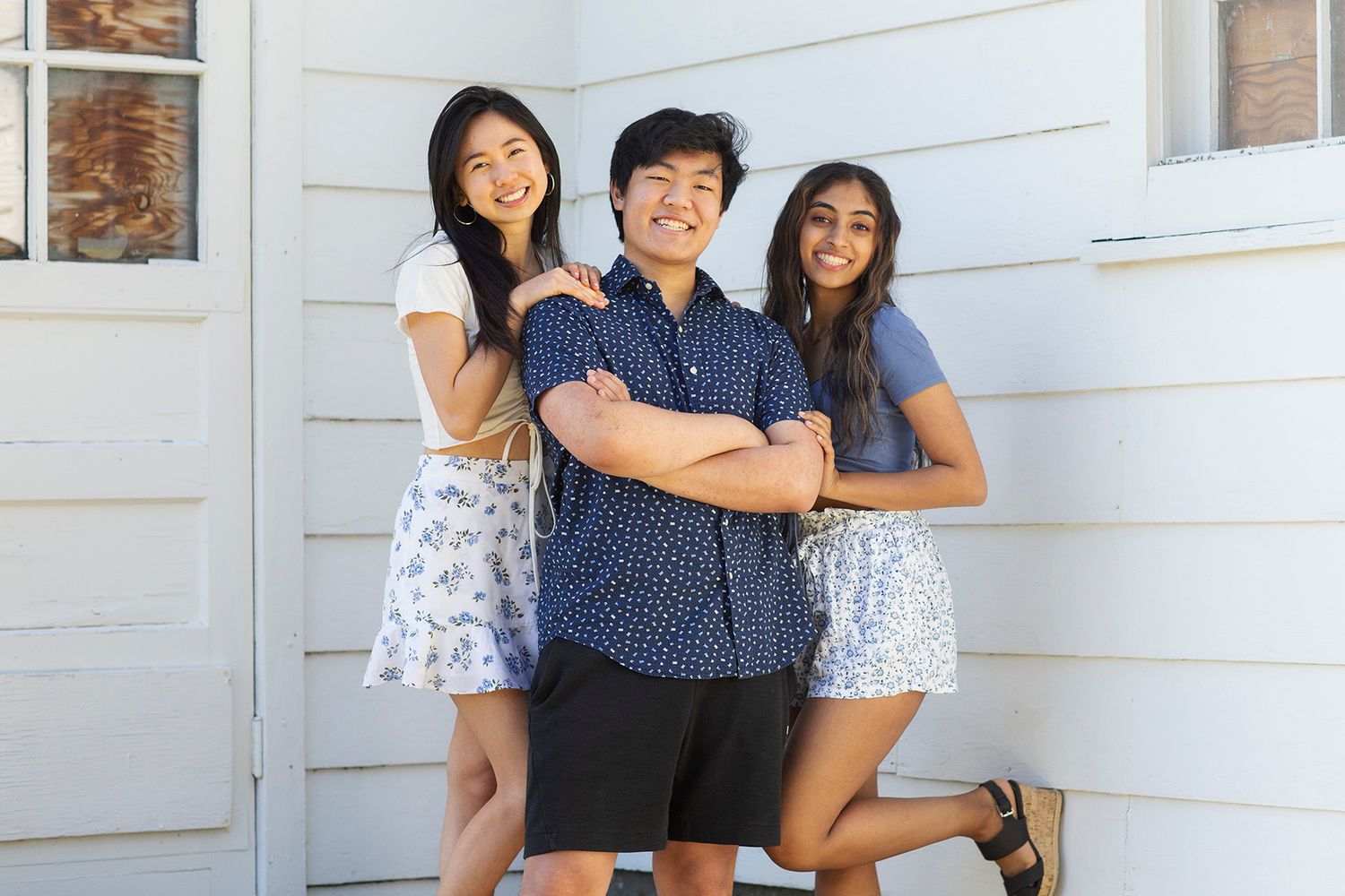 Three smiling teenagers posing together outdoors in front of a white house, wearing casual summer outfits.