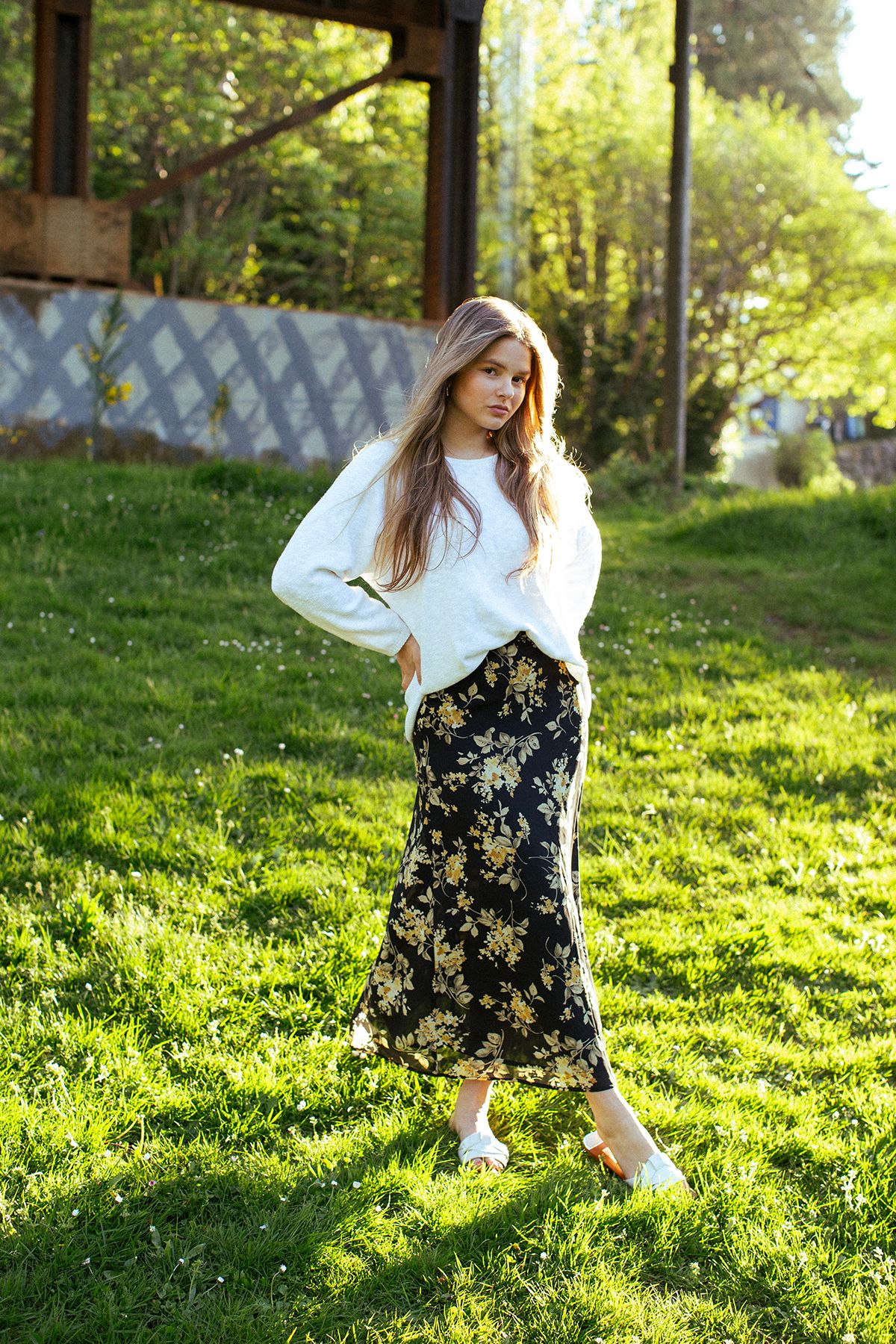Young woman in white sweater and floral midi skirt posing outdoors on green grass in natural sunlight.