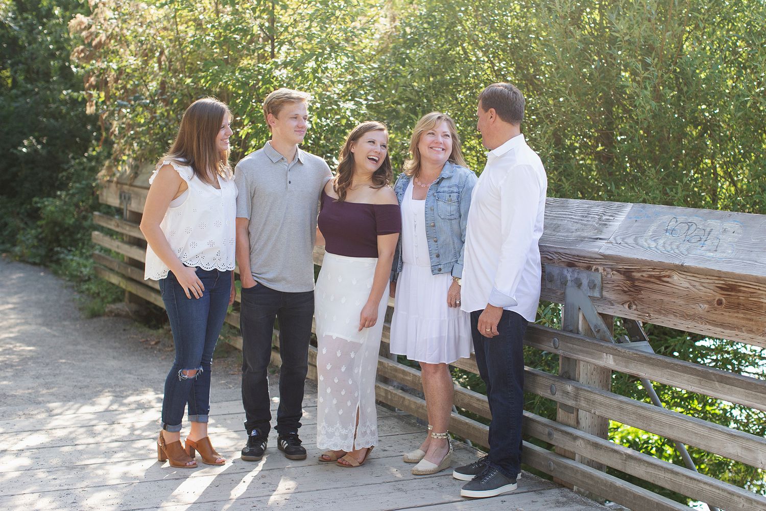 Five people smiling and laughing together on a wooden bridge surrounded by lush green trees on a sunny day.