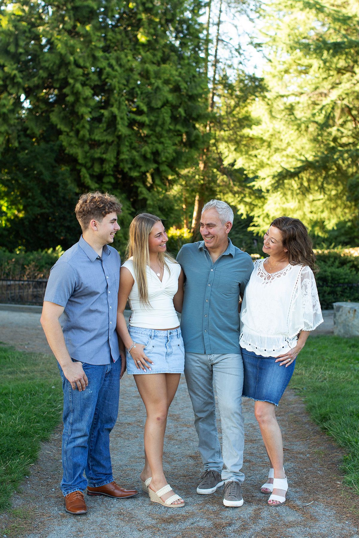 A happy family of four poses together on a park path surrounded by lush green trees on a sunny day.