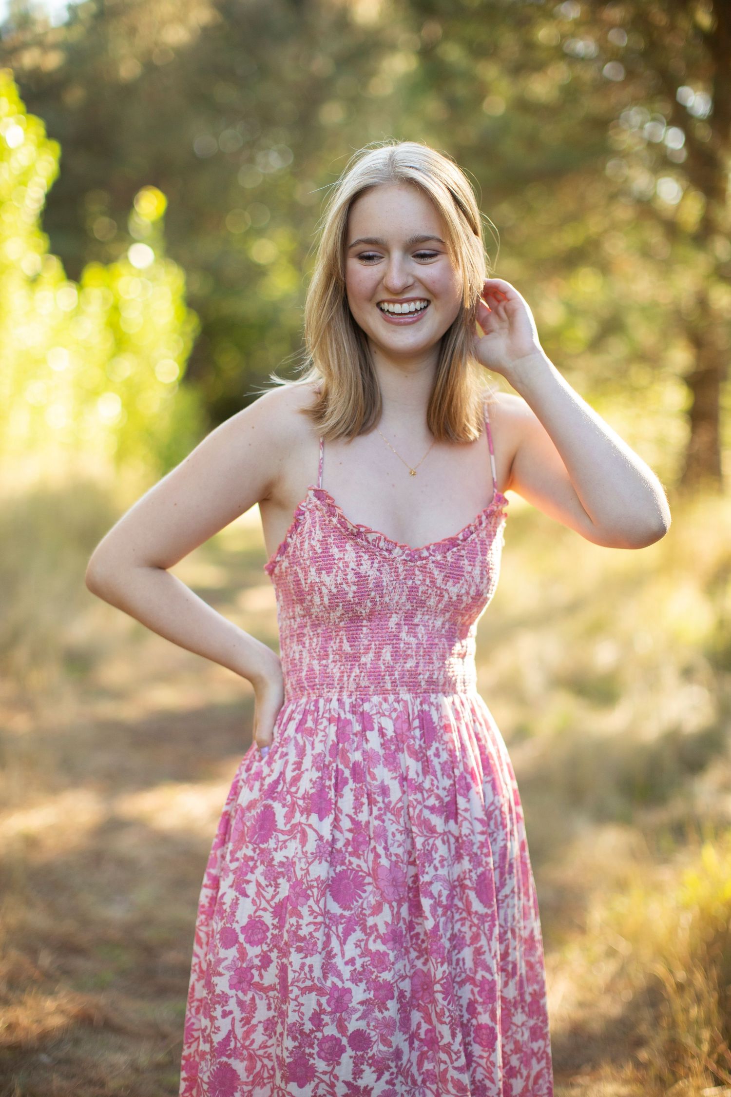 Laughing blonde woman in pink floral sundress standing on sunny outdoor path.