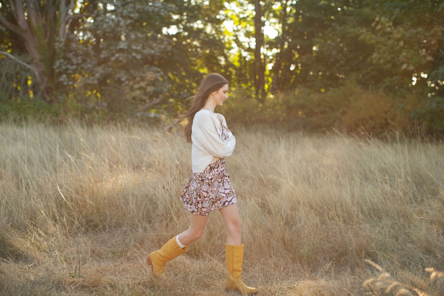 Young woman in floral skirt and cream sweater strolls through golden grass field wearing tan boots.